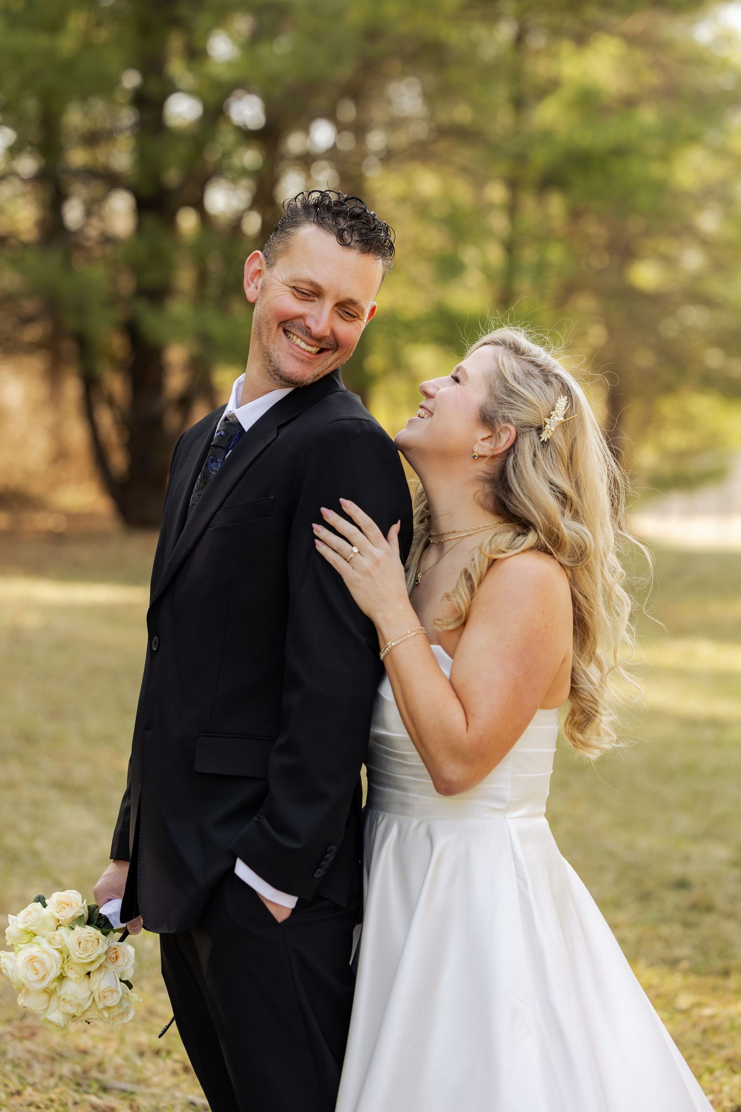 A smiling bride and groom pose outdoors in wedding attire, she holds his arm laughing while he holds white roses.