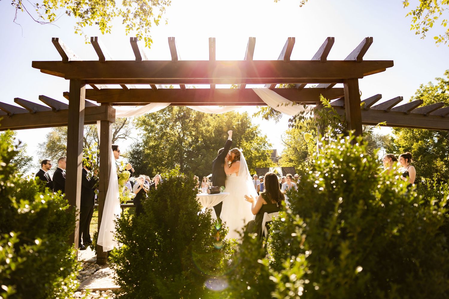 Bride and groom walking through pergola arch in sunlit garden after outdoor wedding ceremony.