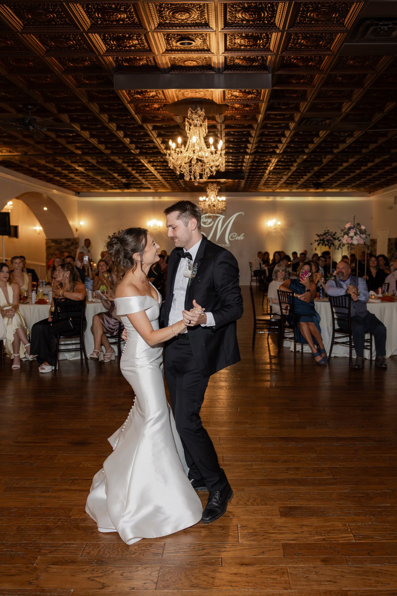 Bride and groom sharing first dance under elegant chandelier in ornate ballroom.