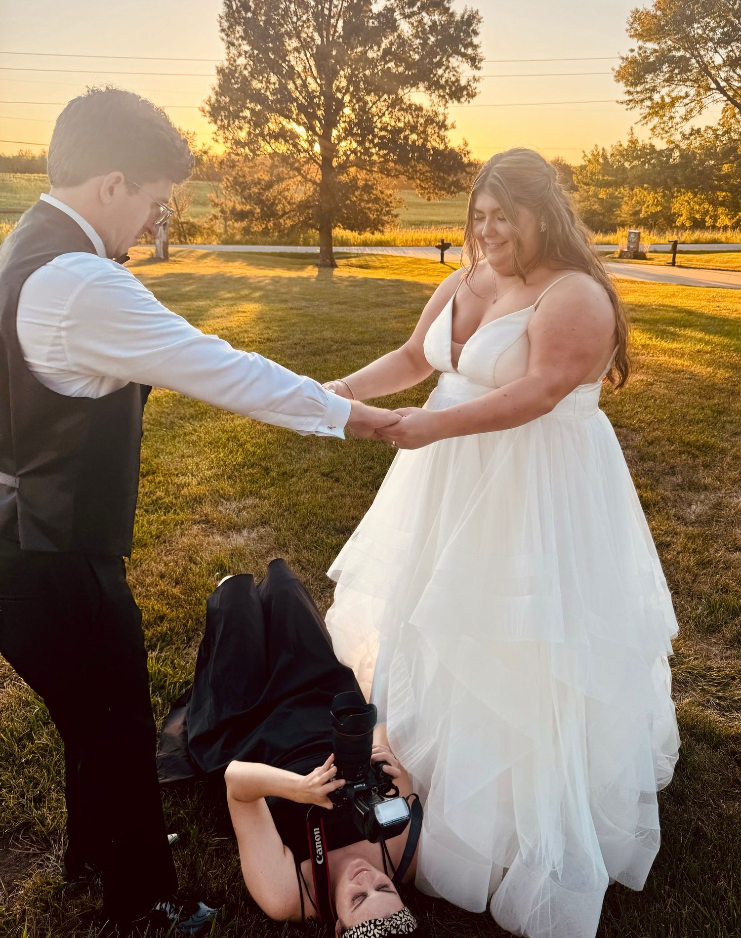 Photographer lying on ground capturing wedding couple holding hands at golden sunset outdoors.