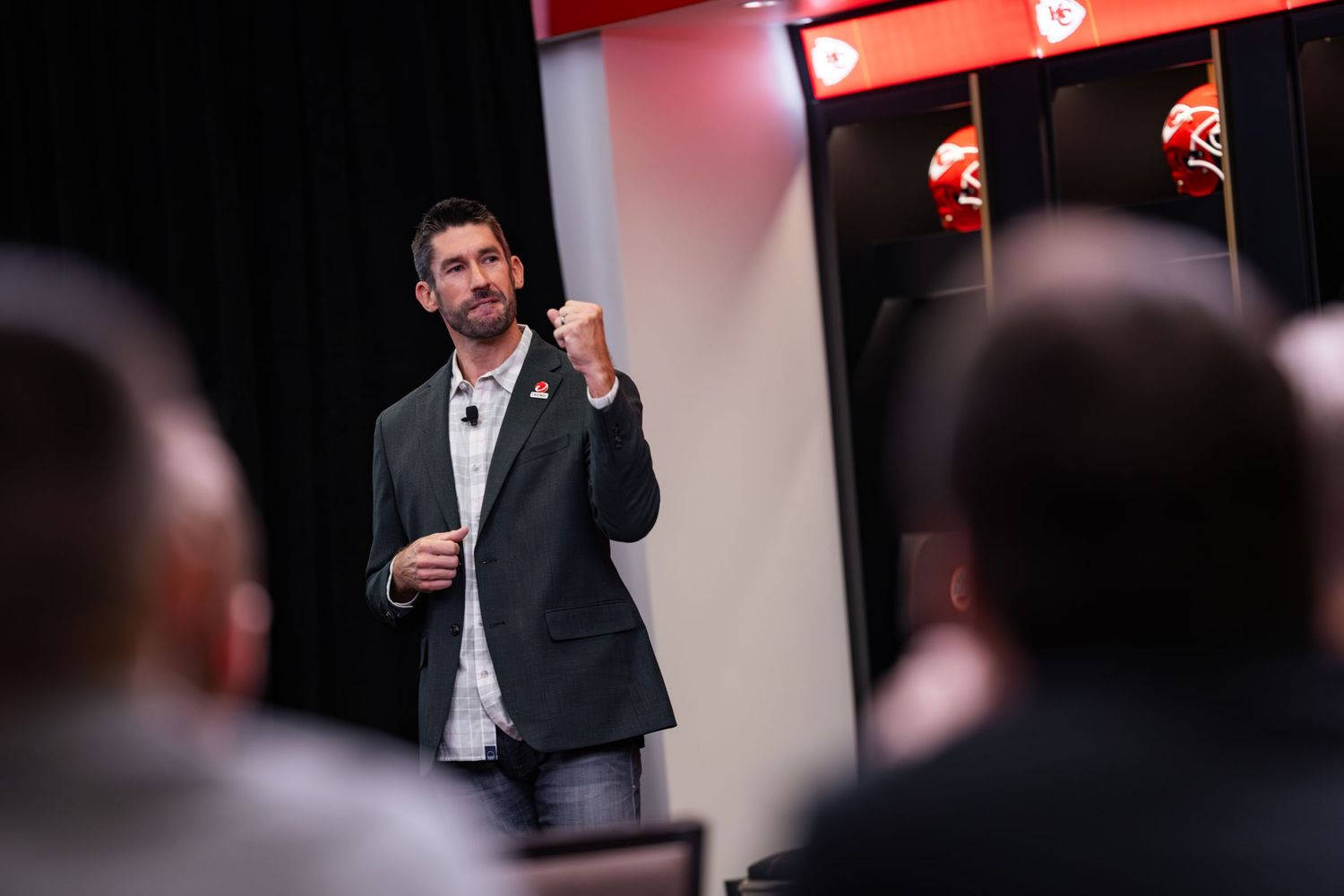 Male speaker presenting on stage in a Kansas City Chiefs locker room venue.