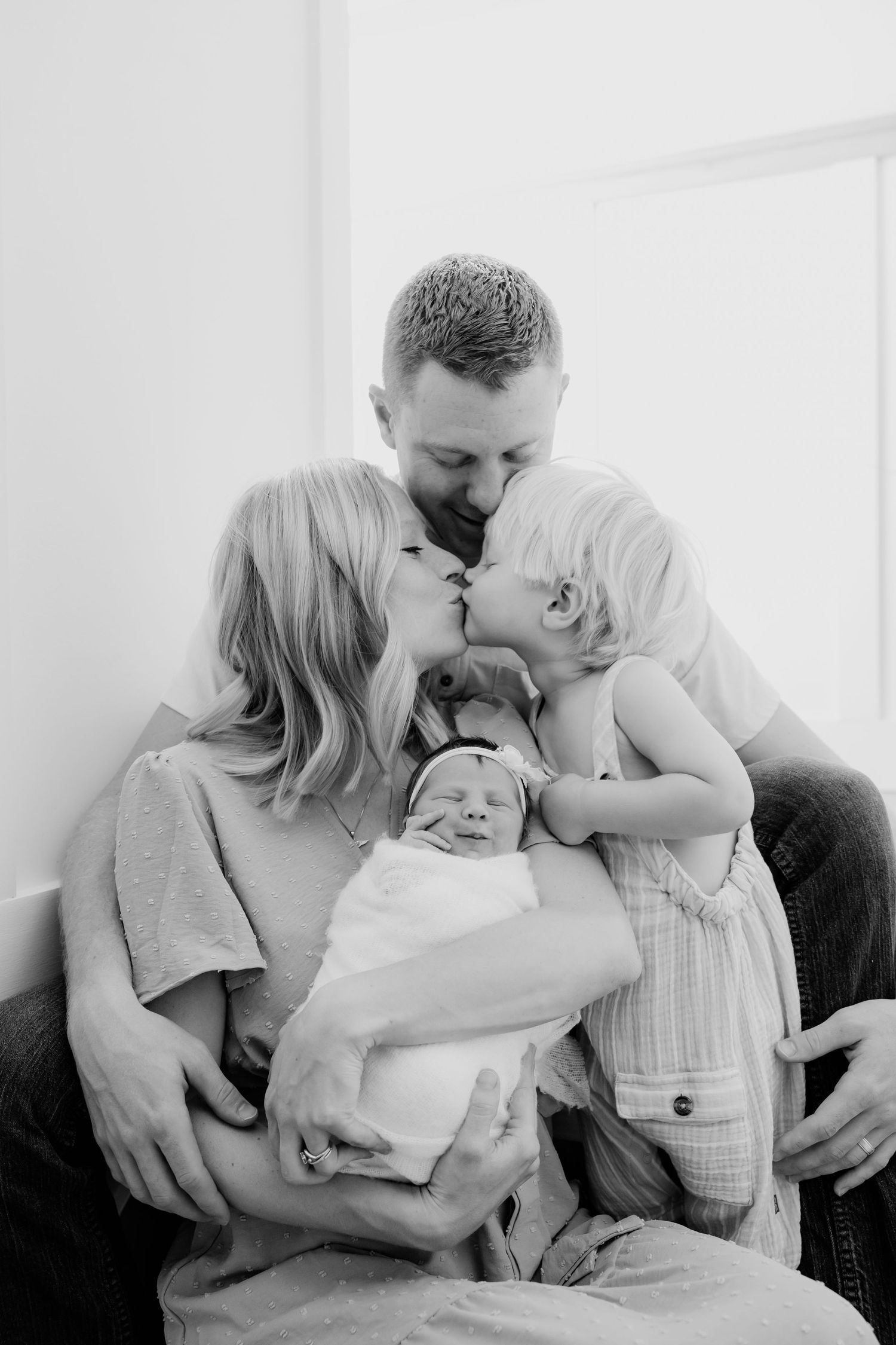 Black and white family photo of parents kissing while holding newborn baby and toddler child.