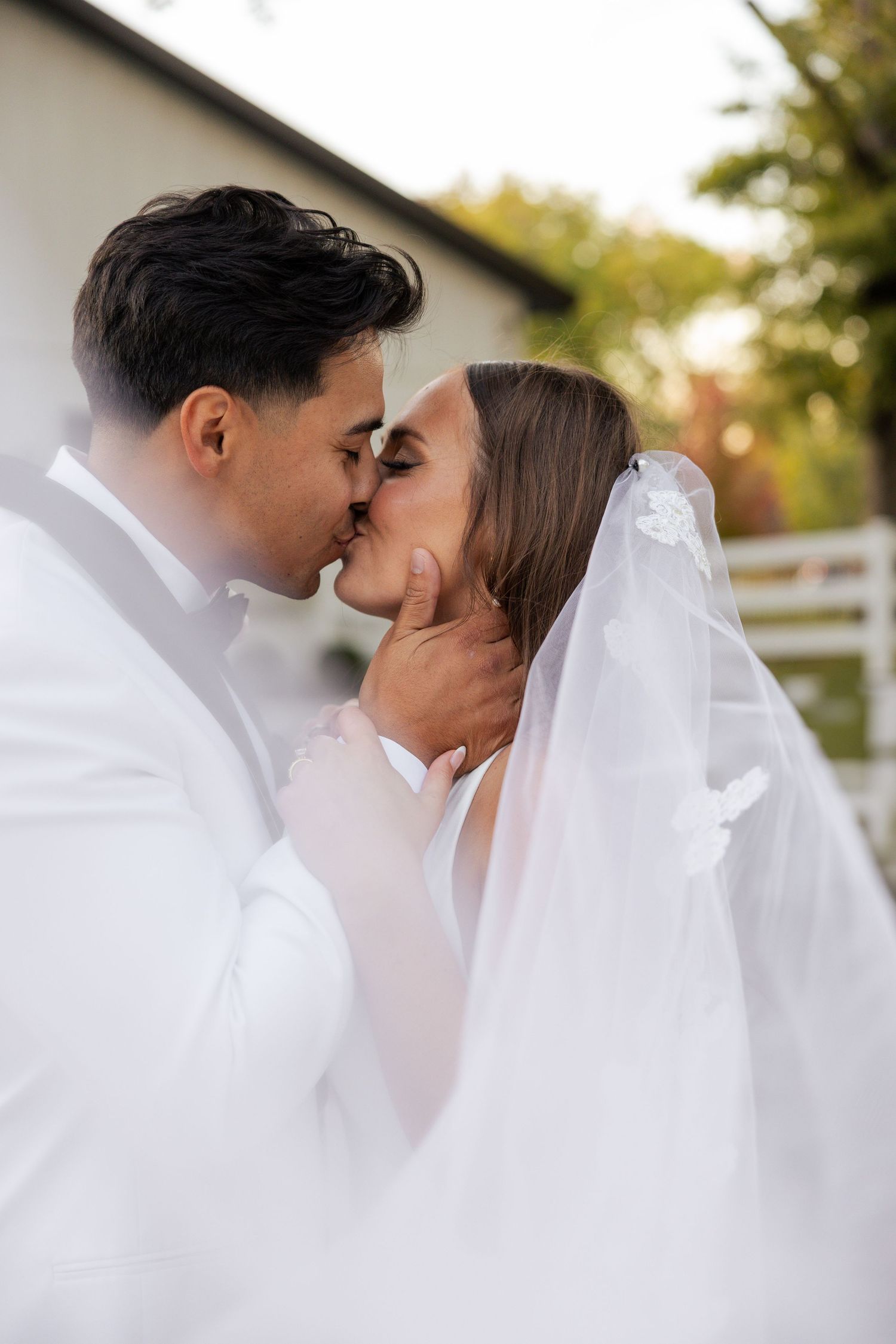 Bride and groom sharing romantic first kiss outdoors, bride wearing floral-trimmed cathedral veil.