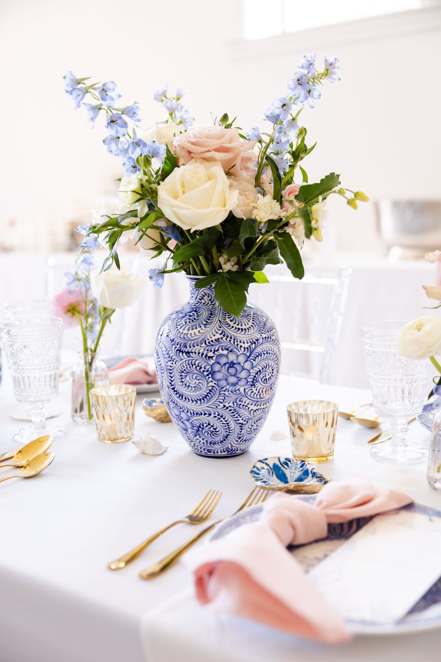 Blue and white vase with roses and delphinium on a white tablecloth with gold cutlery.