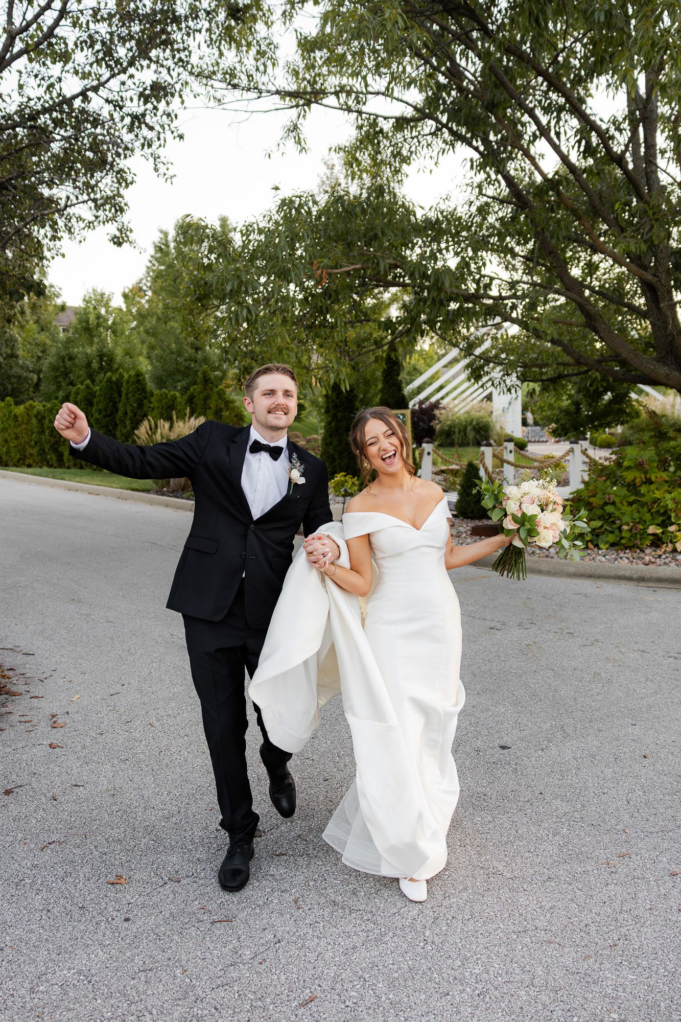 Joyful newlyweds celebrating in a black tuxedo and off-shoulder wedding gown while walking outdoors.