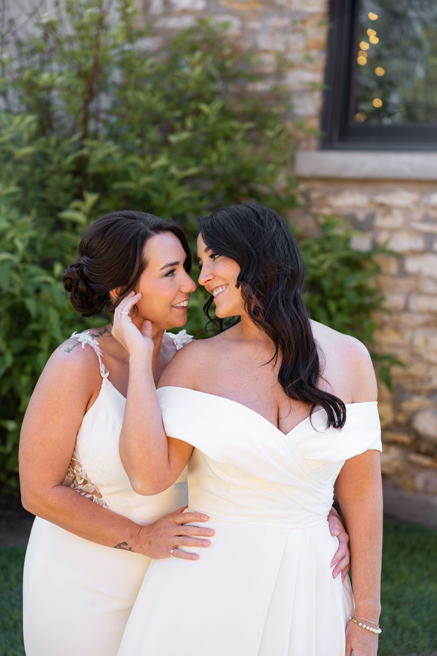 Two brides in elegant white off-shoulder gowns share an intimate moment, smiling at each other outdoors.