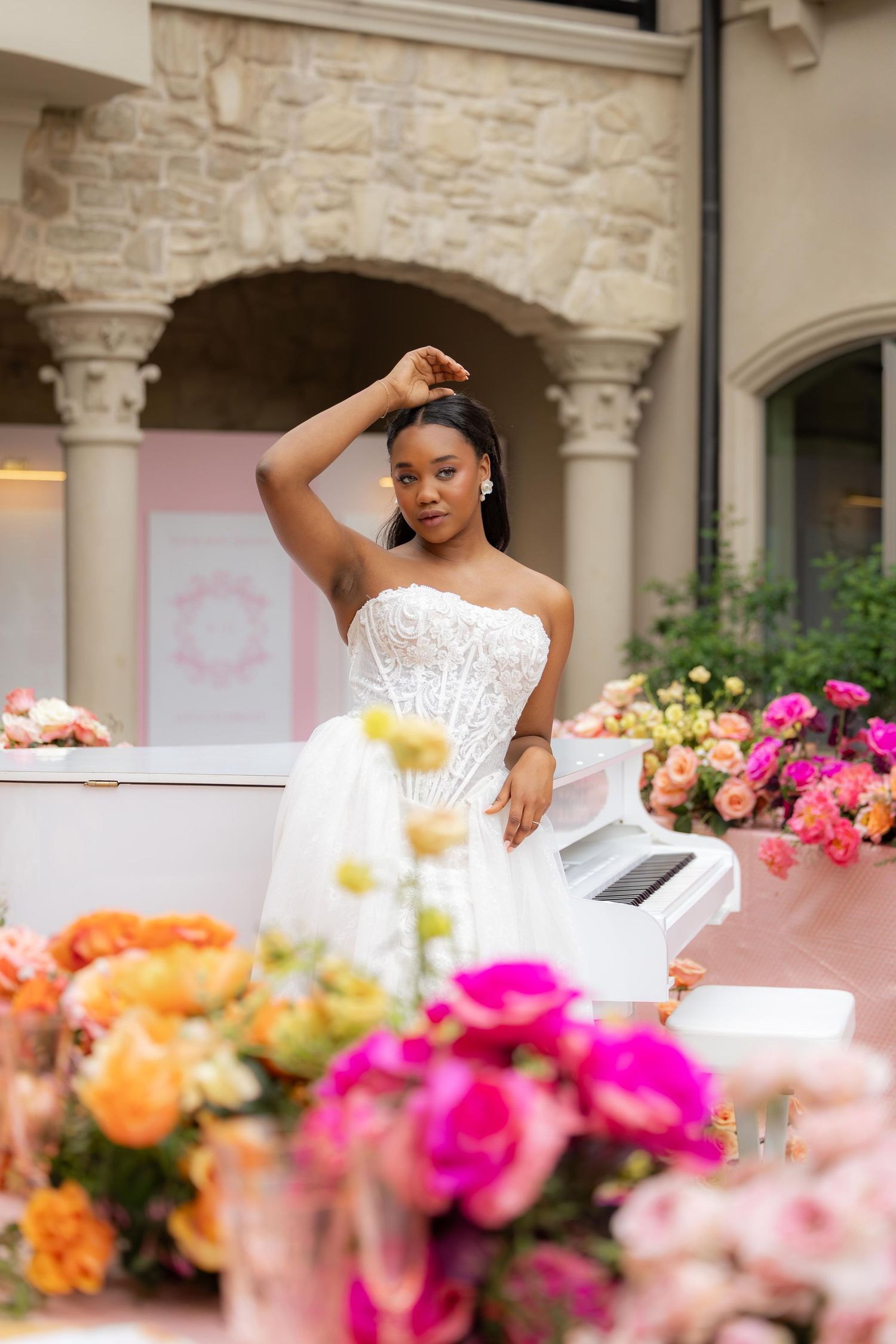 Elegant bride in strapless corset gown poses by white piano surrounded by colorful floral arrangements.