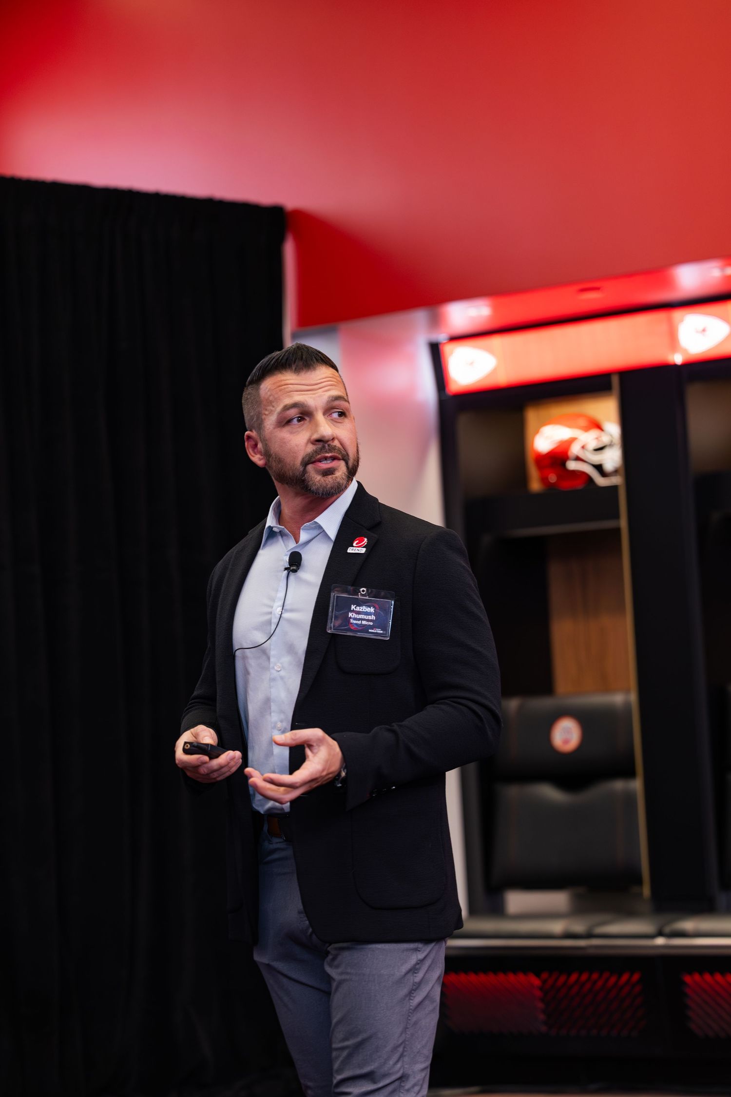 Professional speaker in dark blazer delivering presentation in Kansas City Chiefs facility locker room setting.