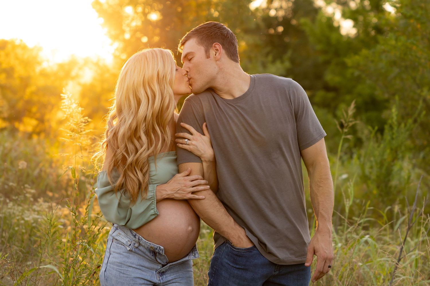 Pregnant couple kissing at golden hour in a field, man embracing woman showing baby bump.