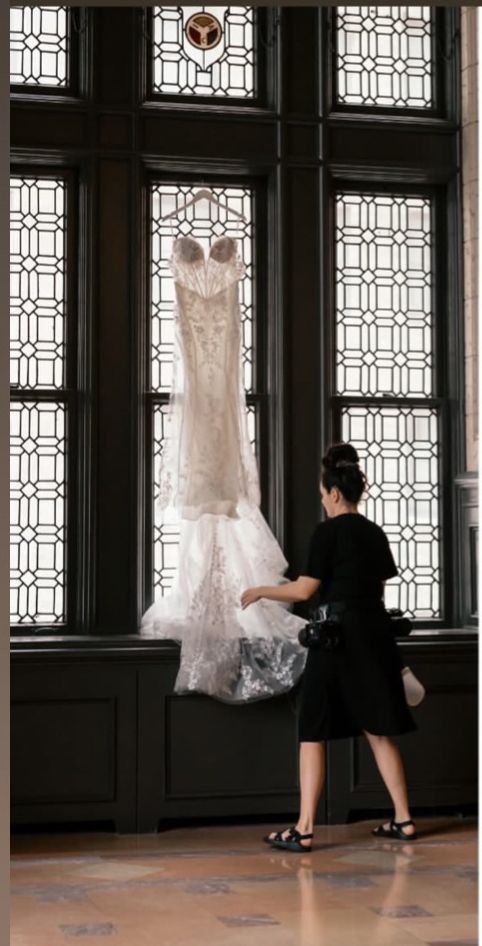 Wedding planner arranging a lace bridal gown hanging in ornate window with geometric glass panes.