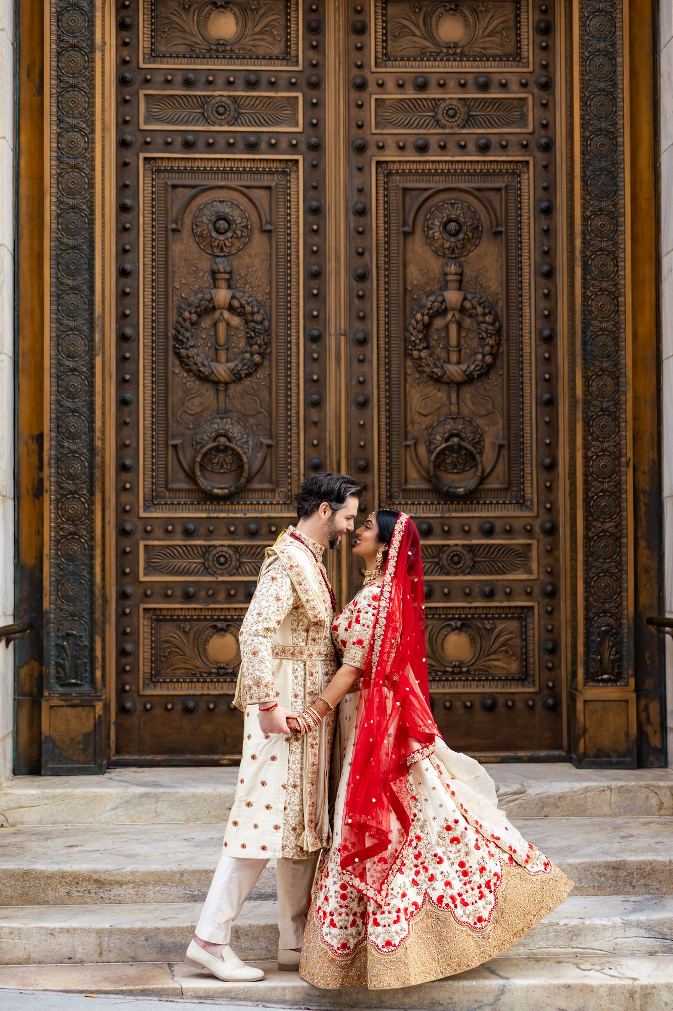 Indian bride and groom in traditional red and gold wedding attire standing before ornate carved wooden doors.