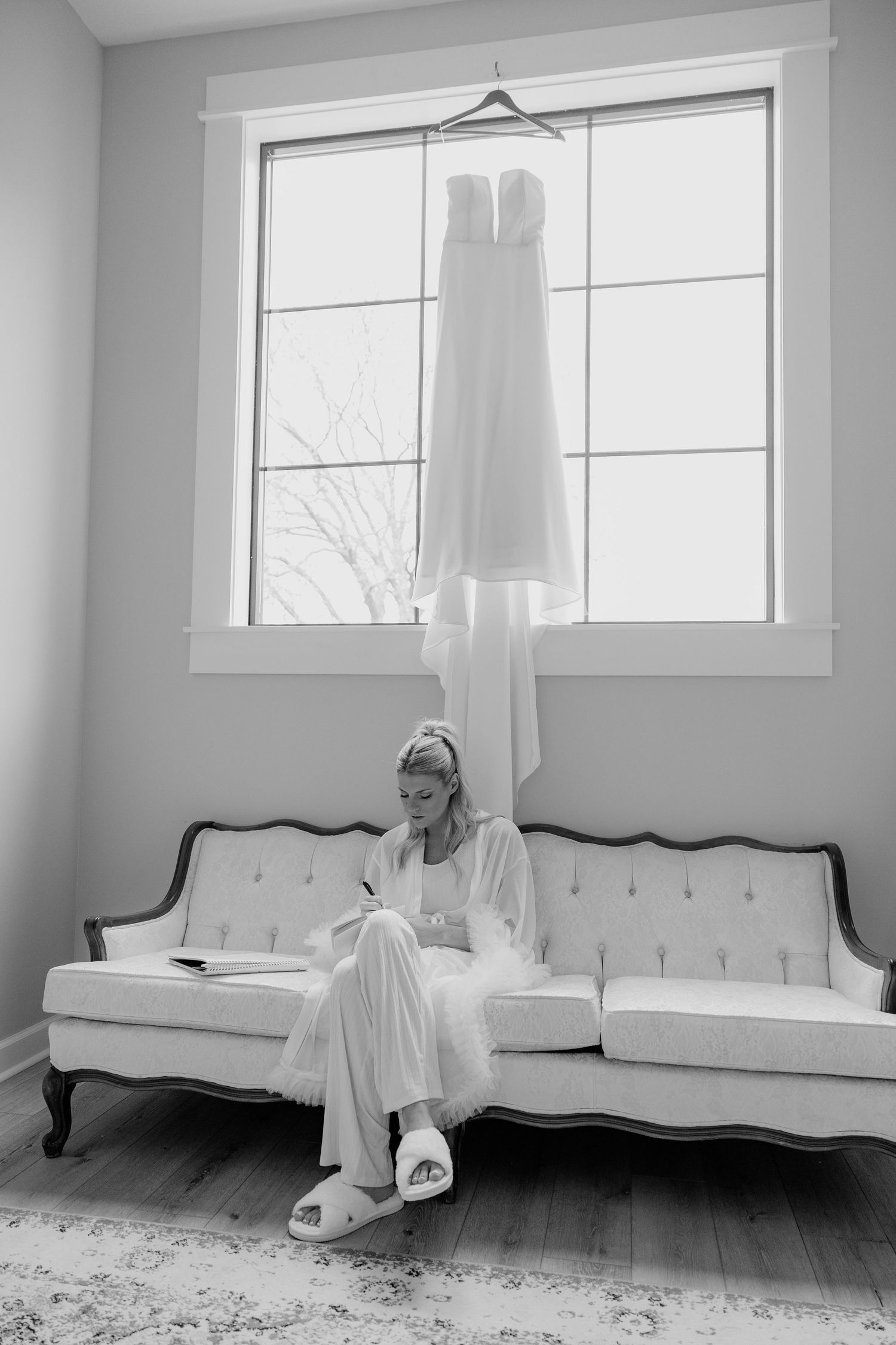 Bride in white robe writing on couch with wedding dress hanging in window above her.