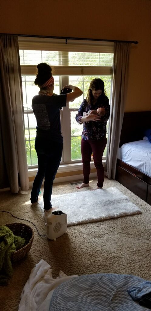 Photographer capturing newborn baby photos as mother holds infant near bright bedroom window.