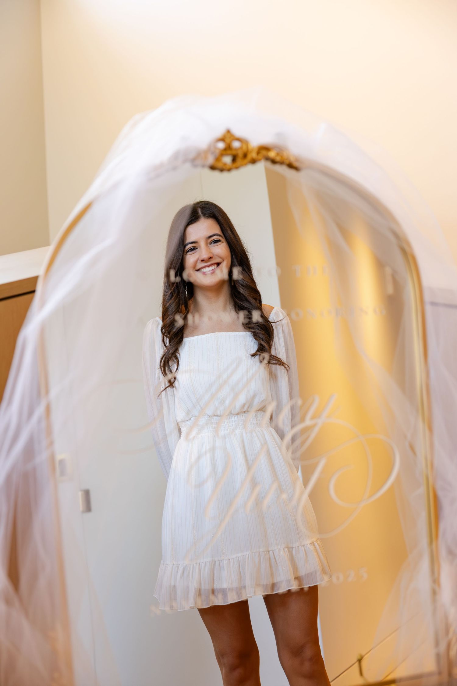 Smiling young woman in white dress holding a flowing veil in a golden-framed mirror.