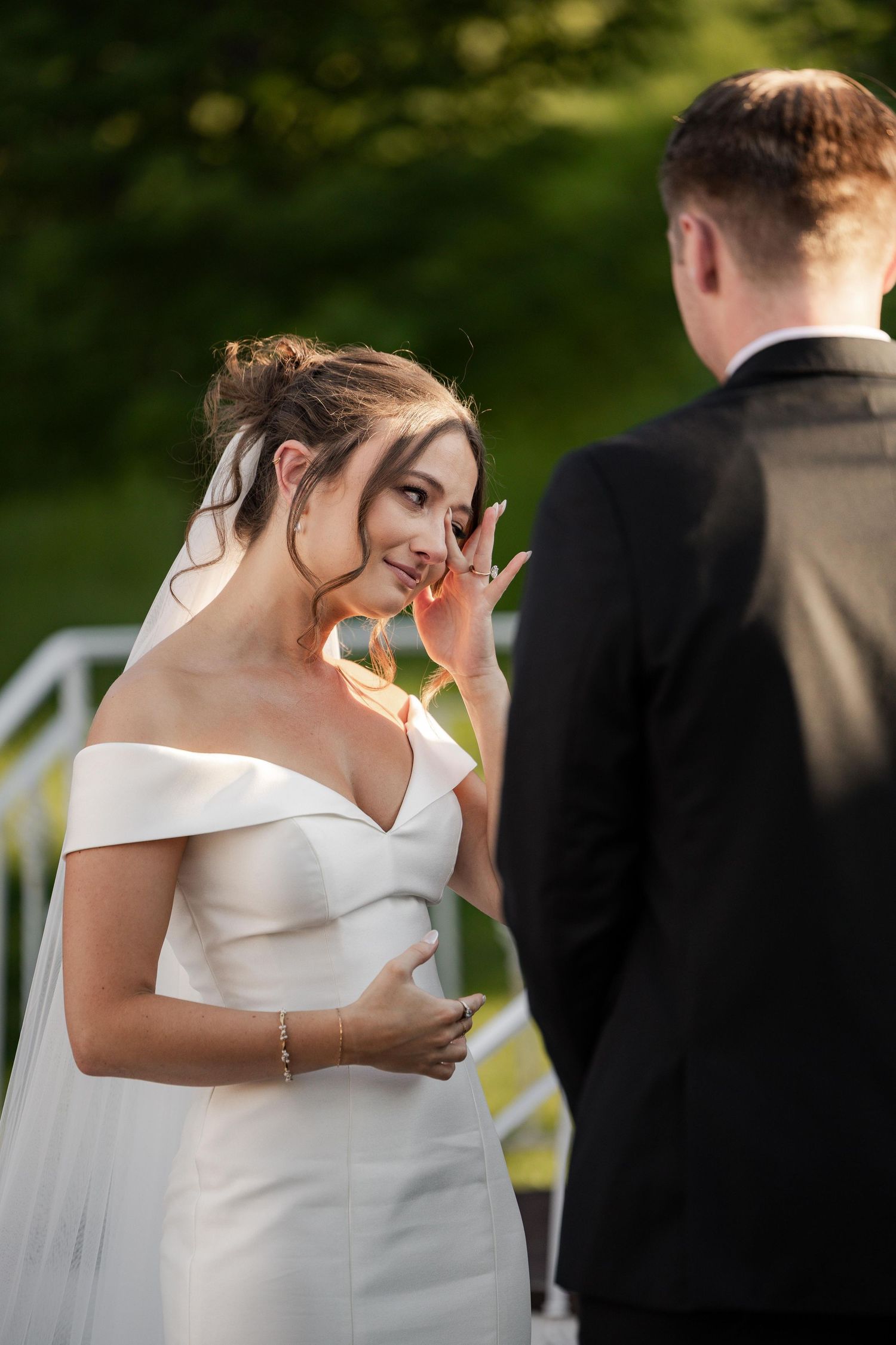Emotional bride in white off-shoulder gown wiping tears during outdoor wedding ceremony with groom in black suit.