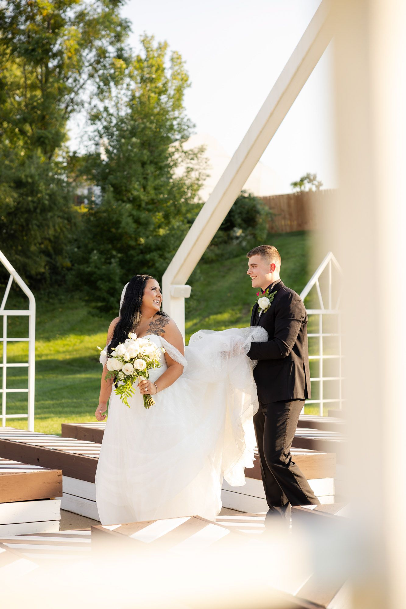 Smiling bride in white gown holds bouquet while groom assists with veil outdoors.
