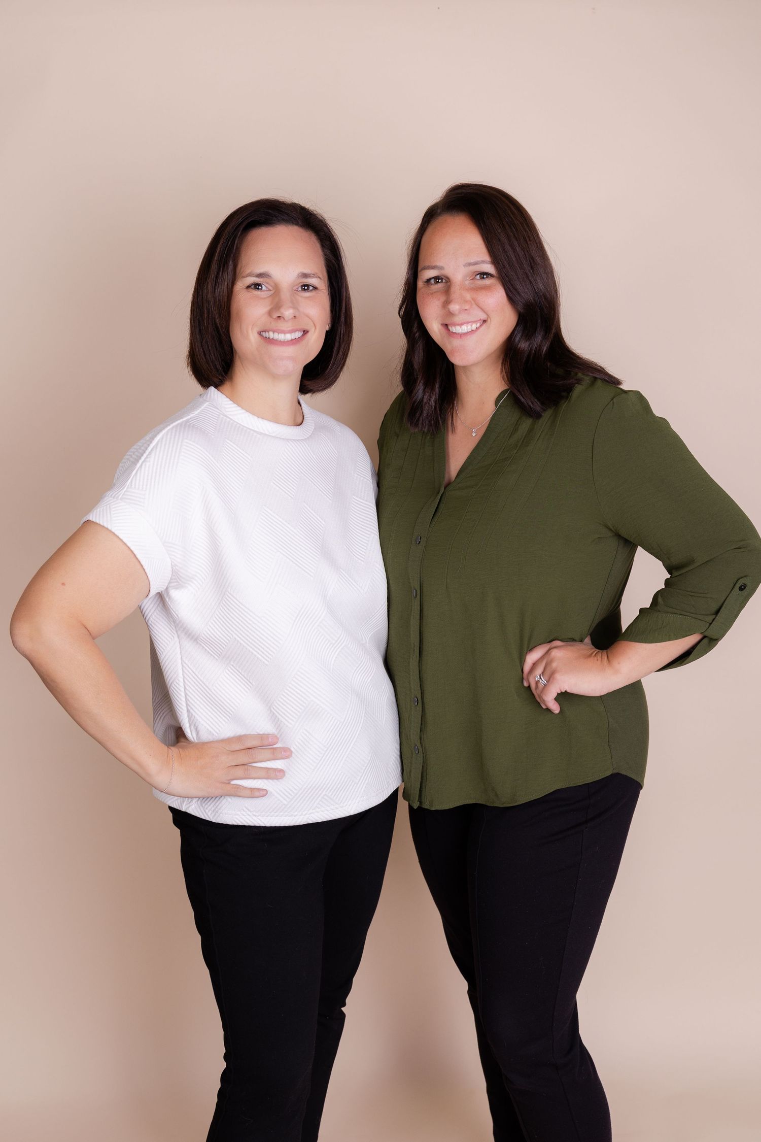 Two smiling women with dark hair pose together, one in a white tee and one in an olive green blouse, against a beige background.