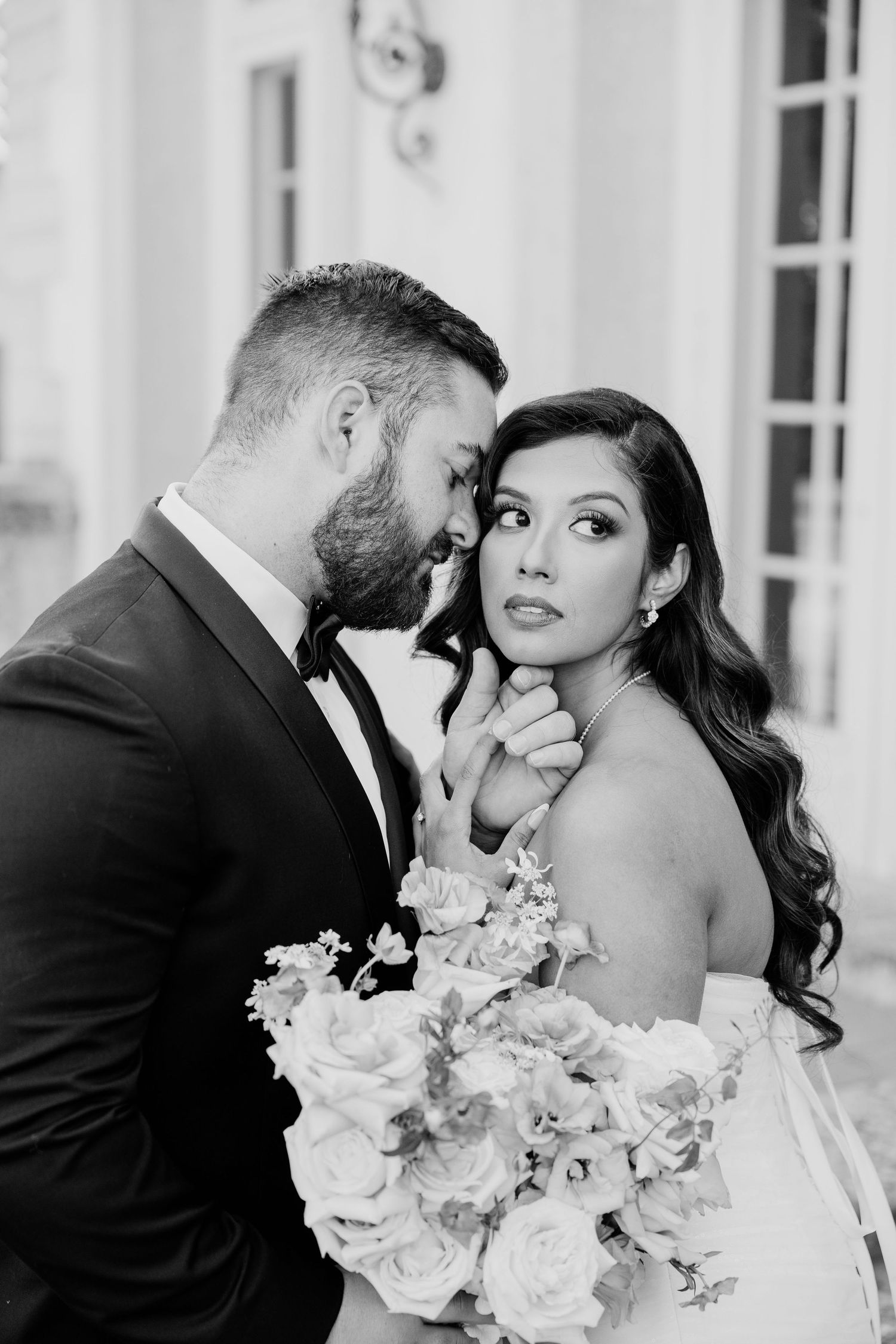 Black and white portrait of bride holding bouquet as groom tenderly holds her chin outdoors.