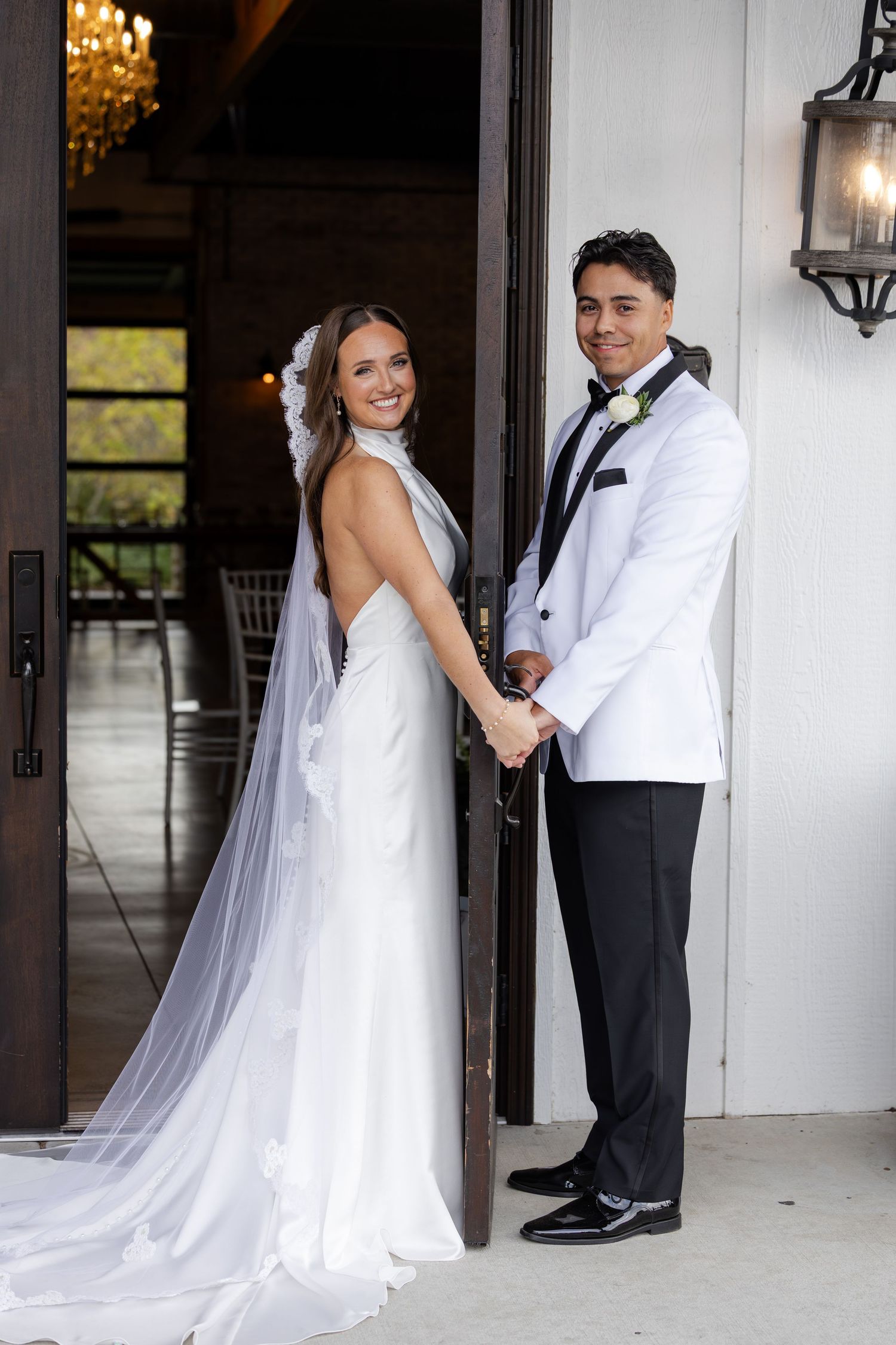 Bride and groom holding hands smiling at camera in doorway, bride wearing backless gown with veil.