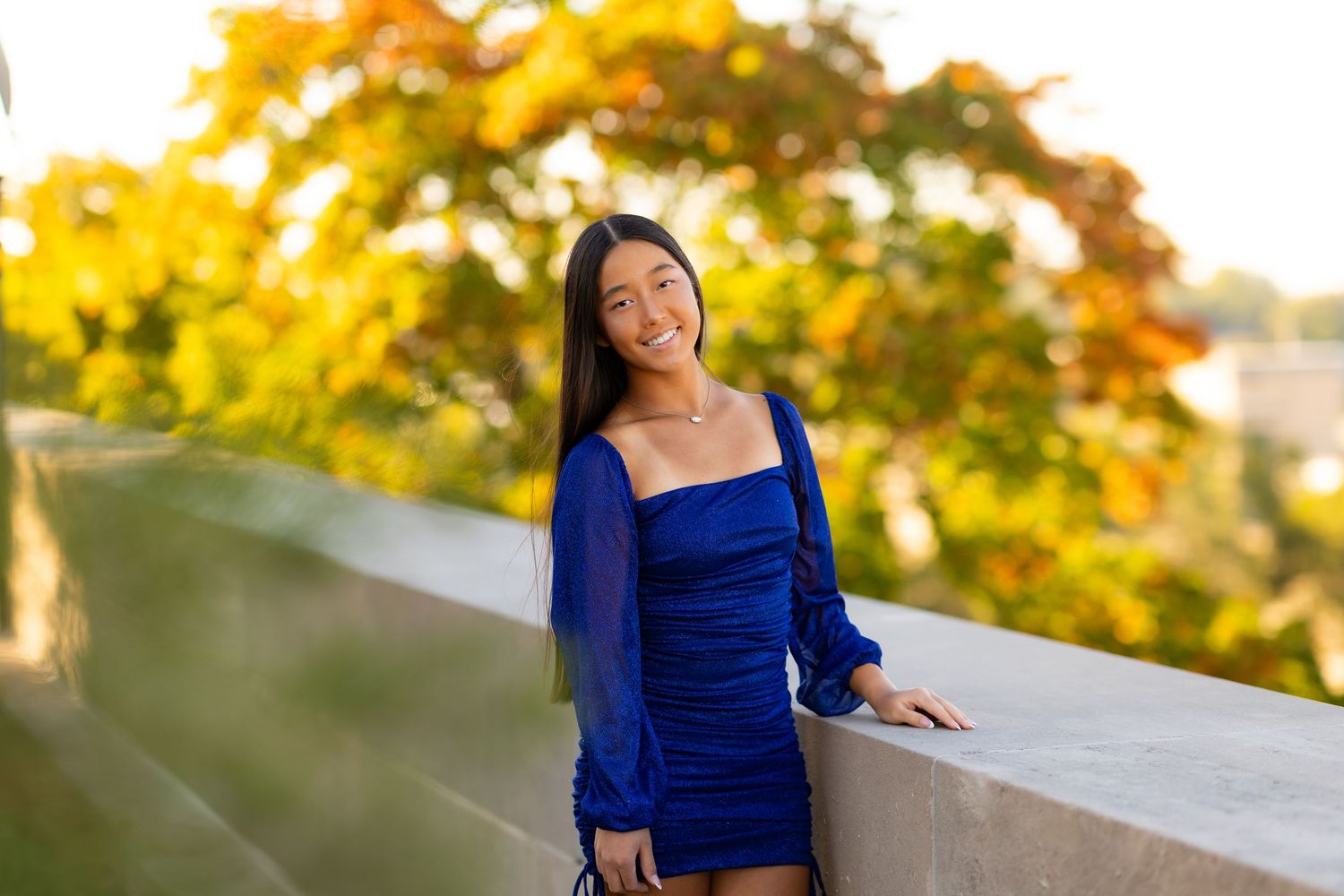 Young woman in blue mini dress smiles while leaning on wall with autumn foliage backdrop.