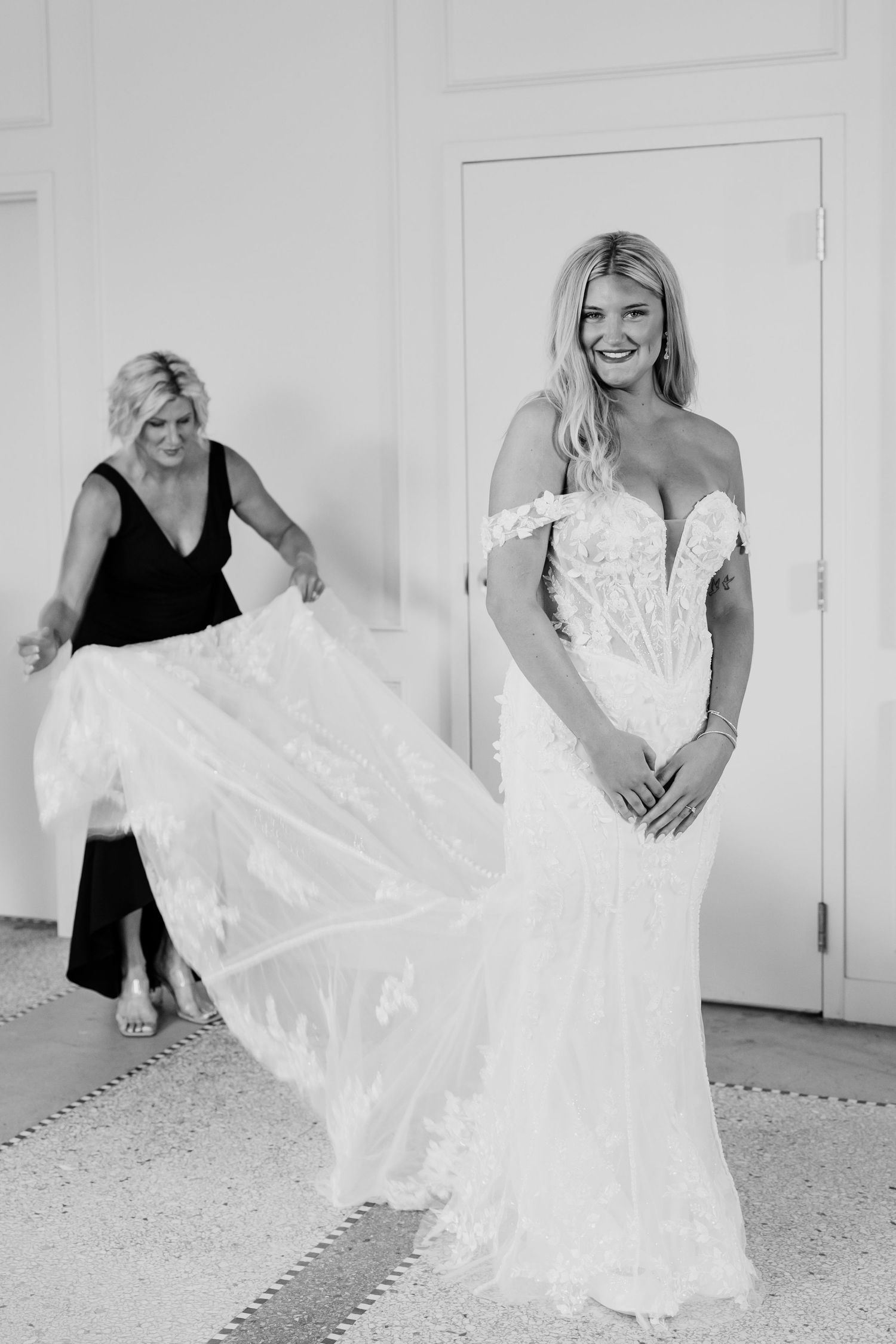 Black and white photo of smiling bride in lace off-shoulder gown while woman adjusts her train indoors.