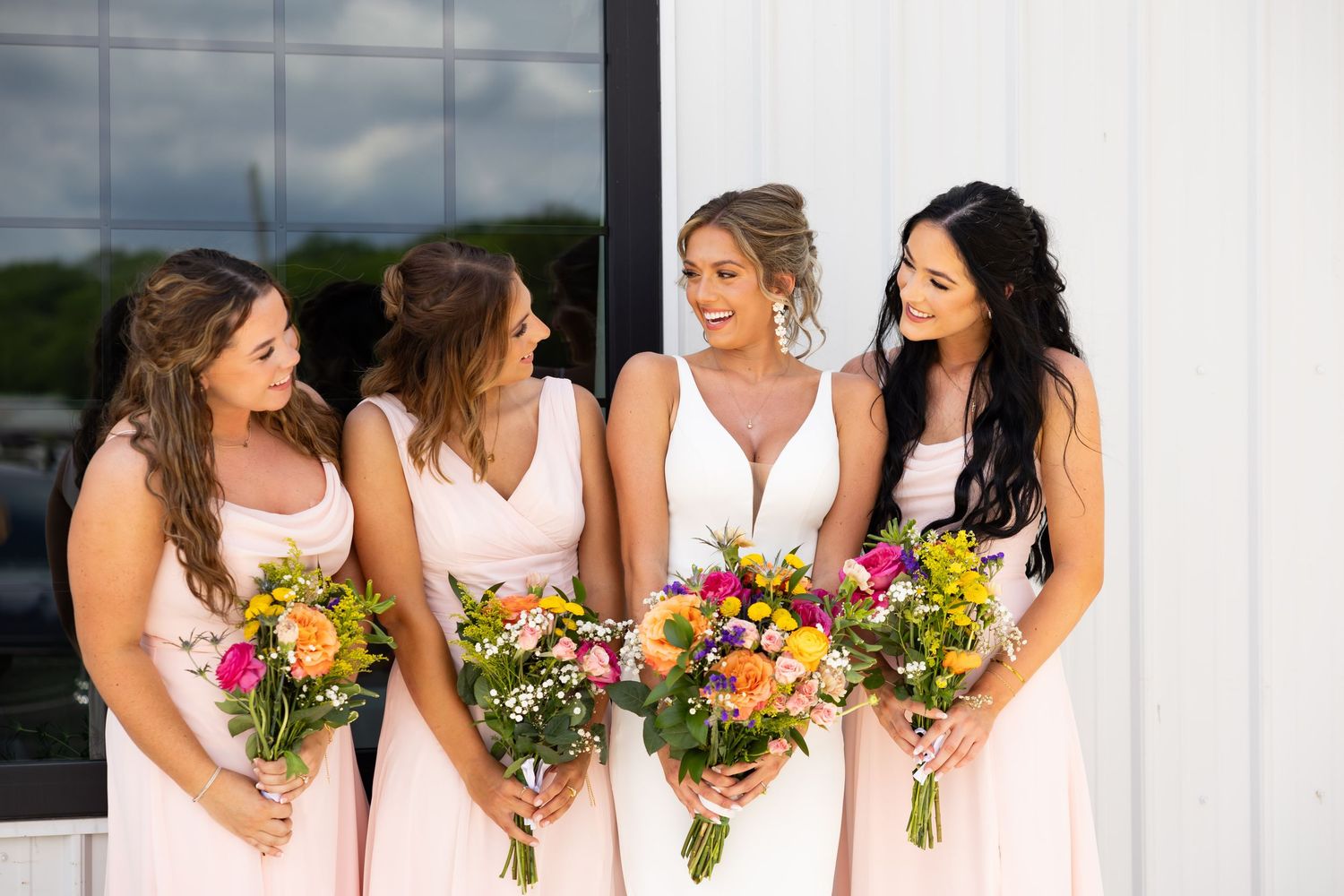 Bride in white gown with three bridesmaids in blush pink dresses holding colorful bouquets.