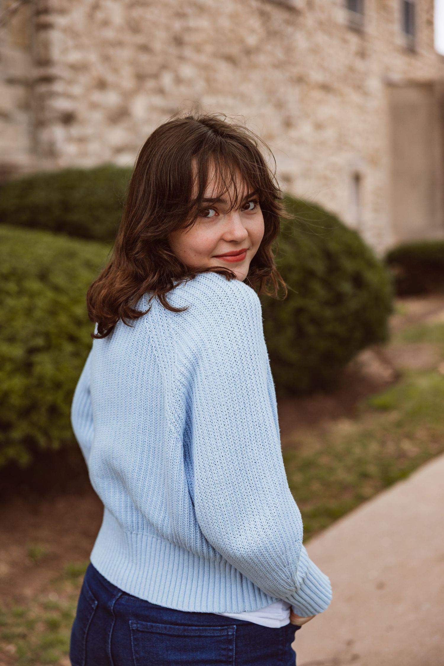 Young brunette woman in light blue knit sweater glancing back over shoulder outdoors near stone building.