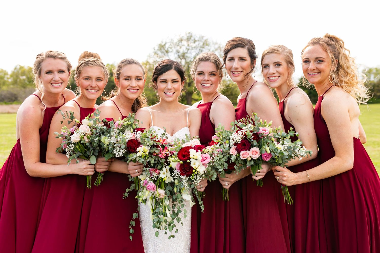 Bride surrounded by eight bridesmaids in matching burgundy gowns holding lush floral bouquets outdoors.