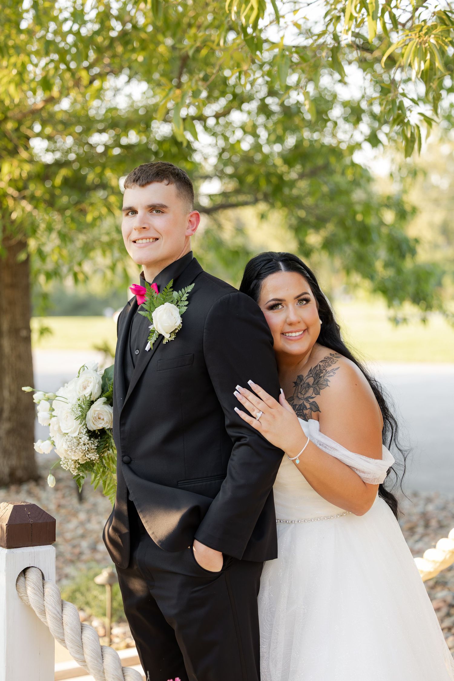 Bride leans on groom's back as couple poses under tree at outdoor wedding venue.