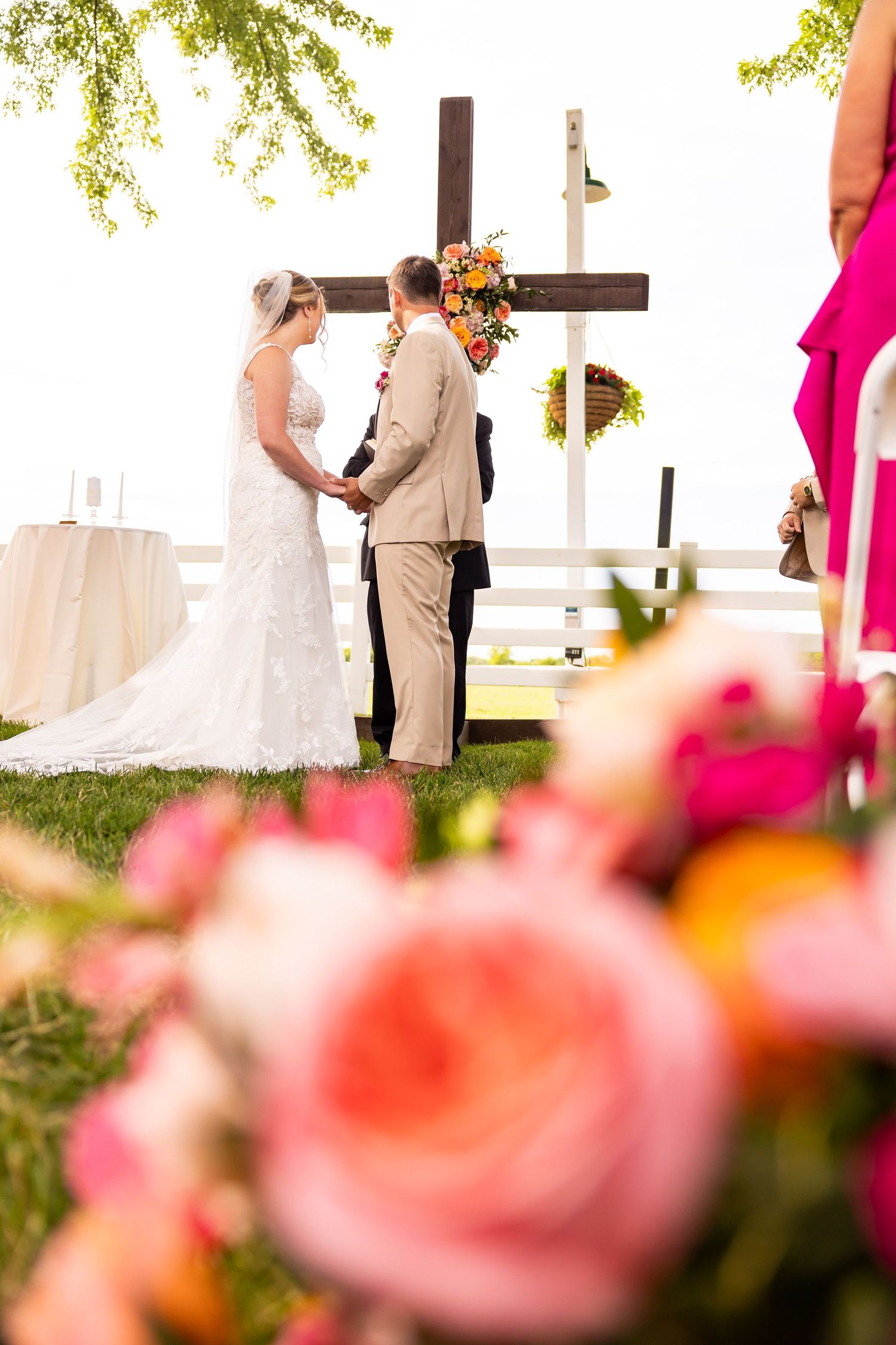 Bride and groom hold hands at outdoor wedding ceremony before a wooden cross with floral decor.
