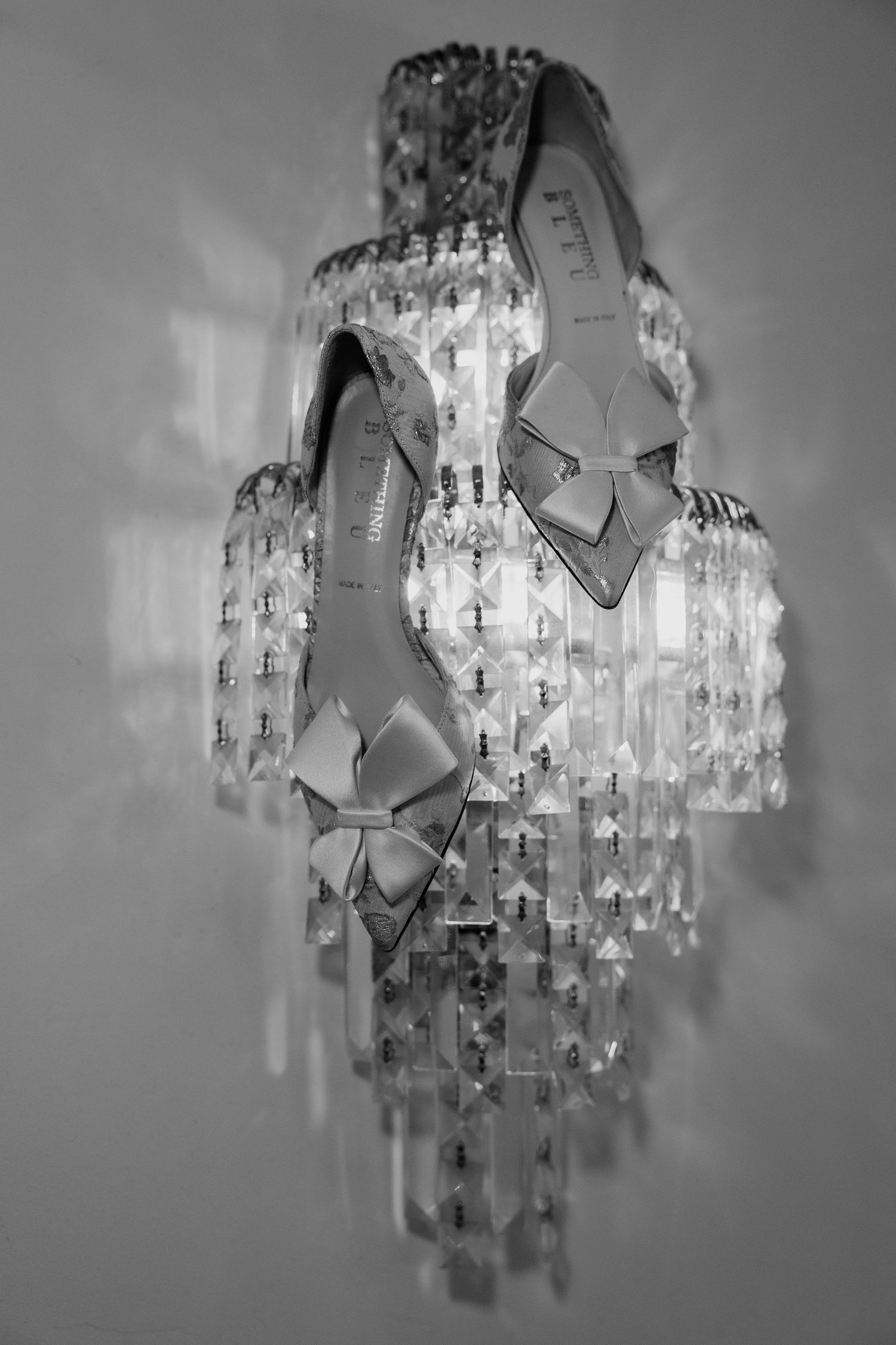 Black and white photo of bridal heels with bow detail displayed on crystal chandelier wall sconce.