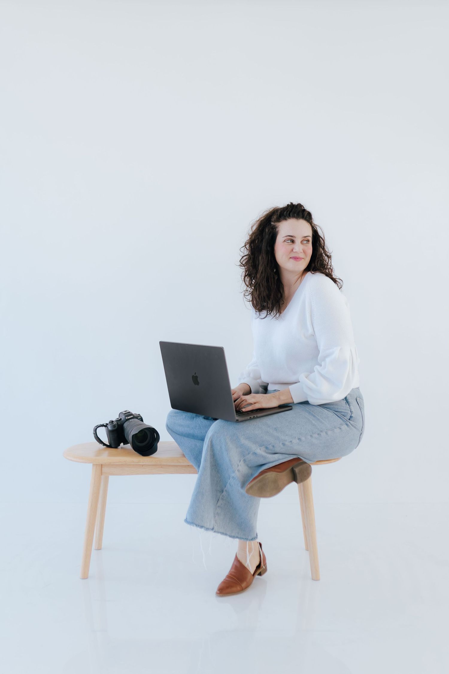Woman in white top seated on wooden bench with laptop open and camera resting beside her.