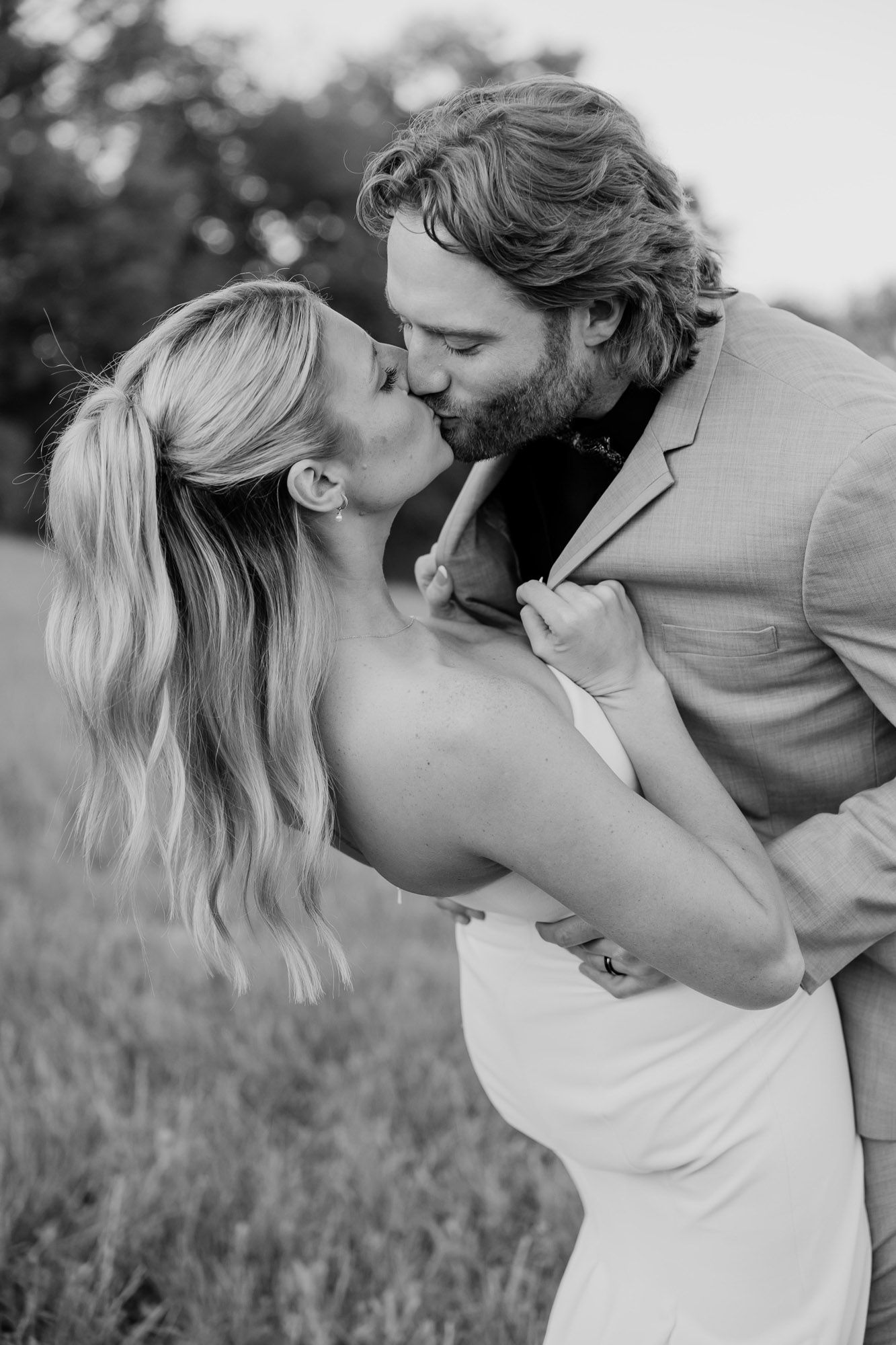 Black and white photo of a couple sharing a passionate kiss in an outdoor field setting.