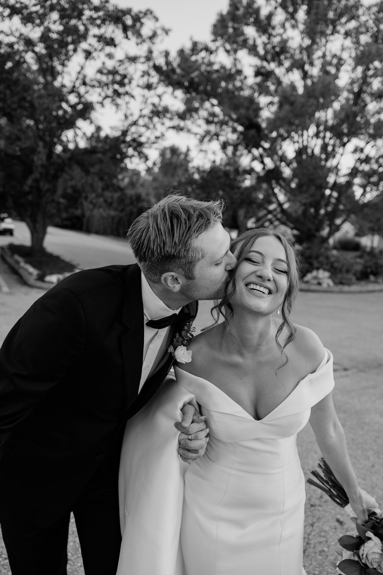 Black and white photo of a groom in a tuxedo kissing his laughing bride on the cheek outdoors.