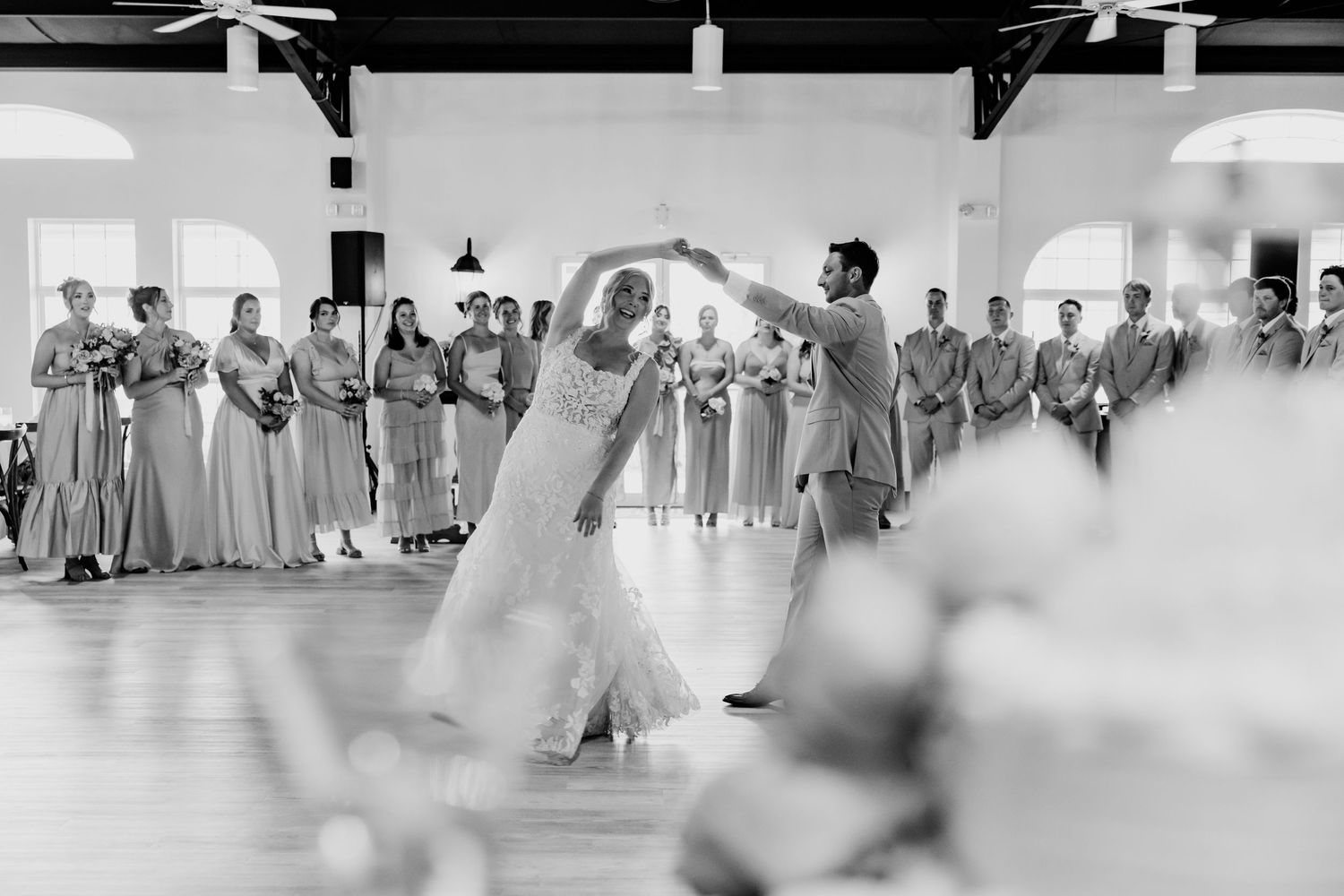 Bride and groom share first dance in black and white photo while wedding party watches indoors.