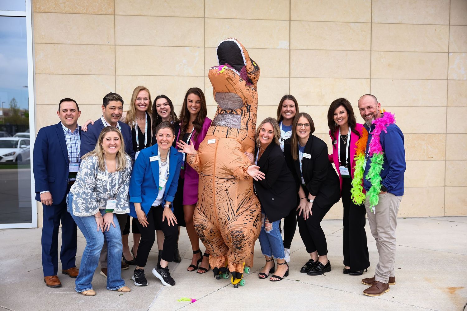 Group of smiling professionals posing outdoors with person in inflatable T-Rex dinosaur costume at event.