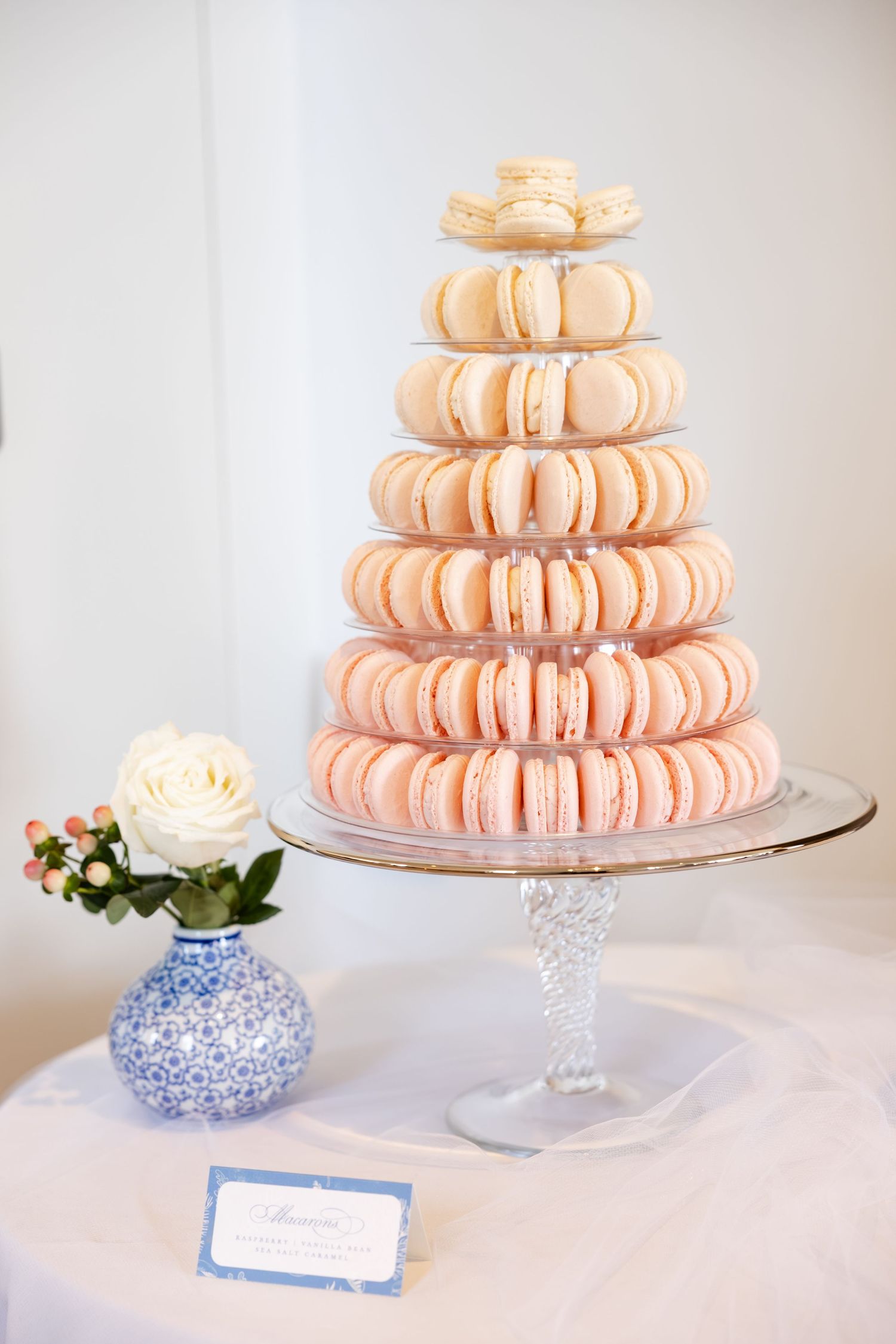Tiered tower of pink and white macarons on crystal stand with blue vase beside it.