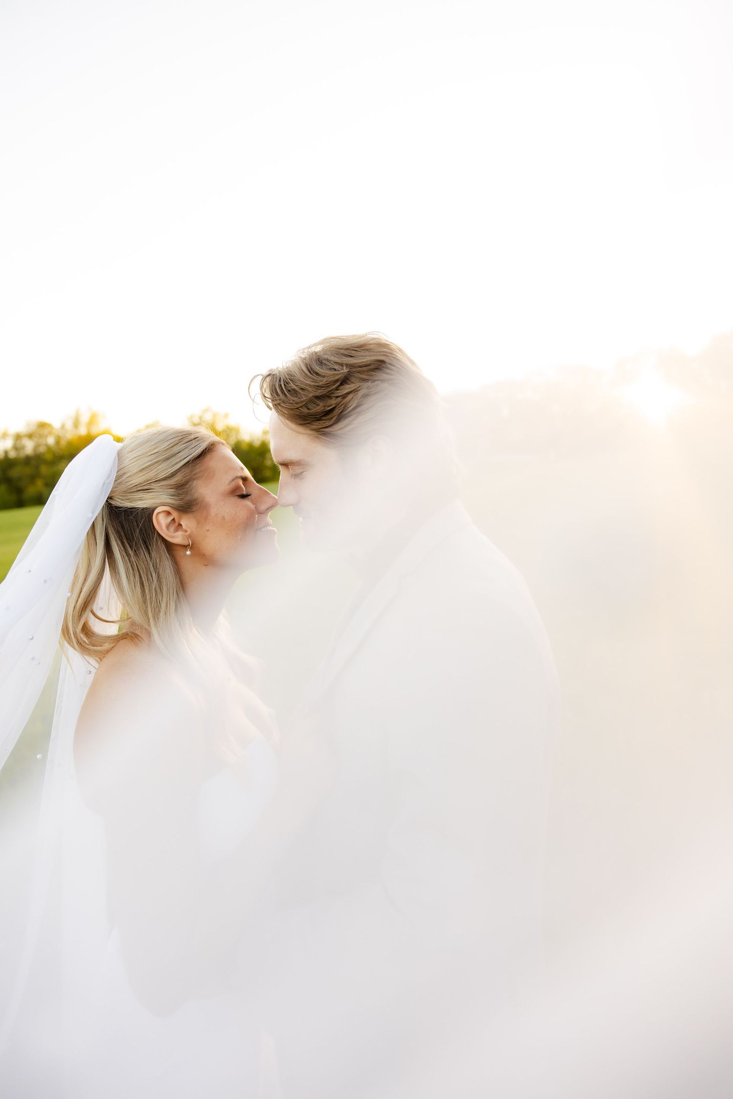 Bride and groom kissing softly as wedding veil billows in bright backlit outdoor setting.