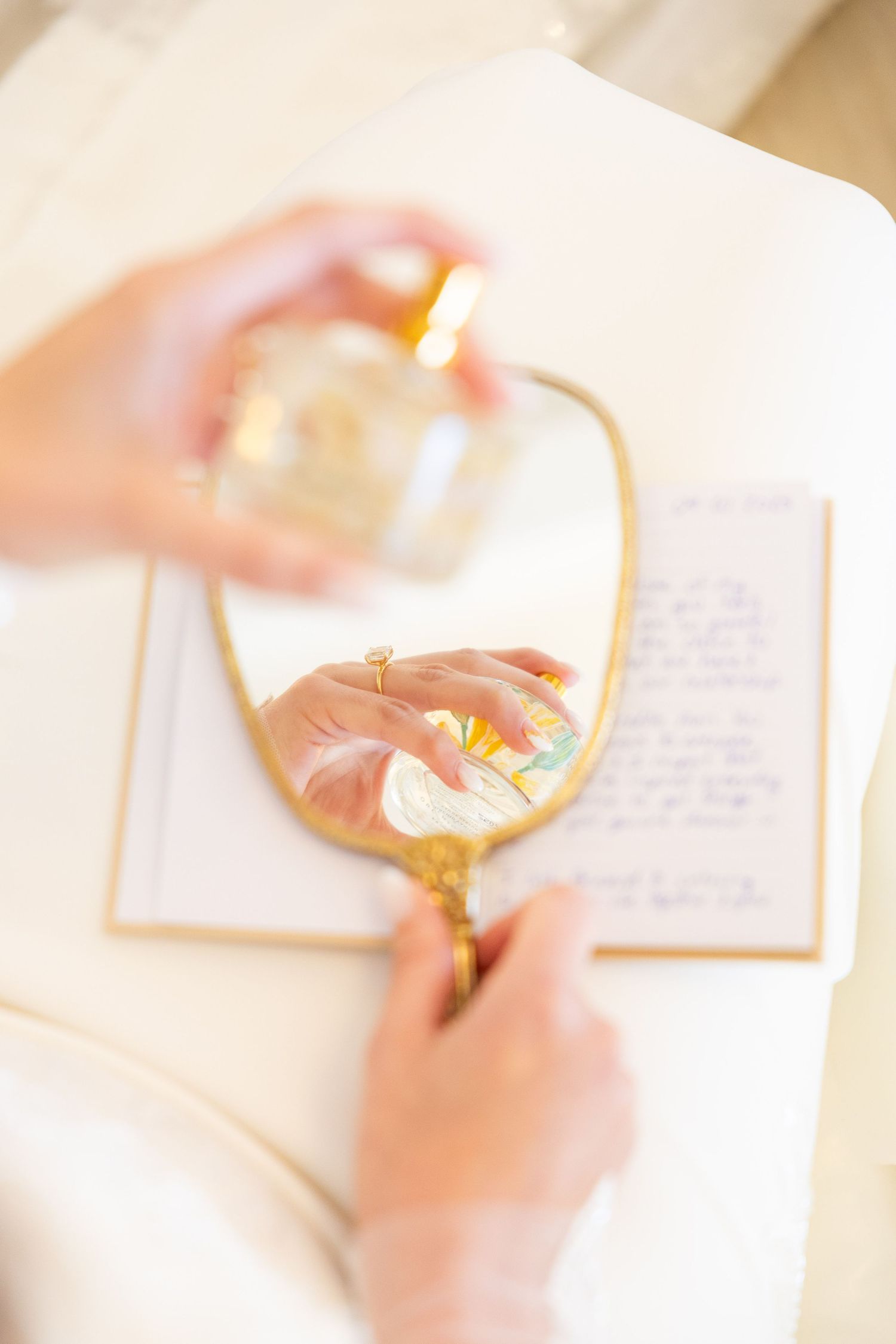 Bride holds perfume bottle reflected in gold oval mirror beside handwritten wedding vows.