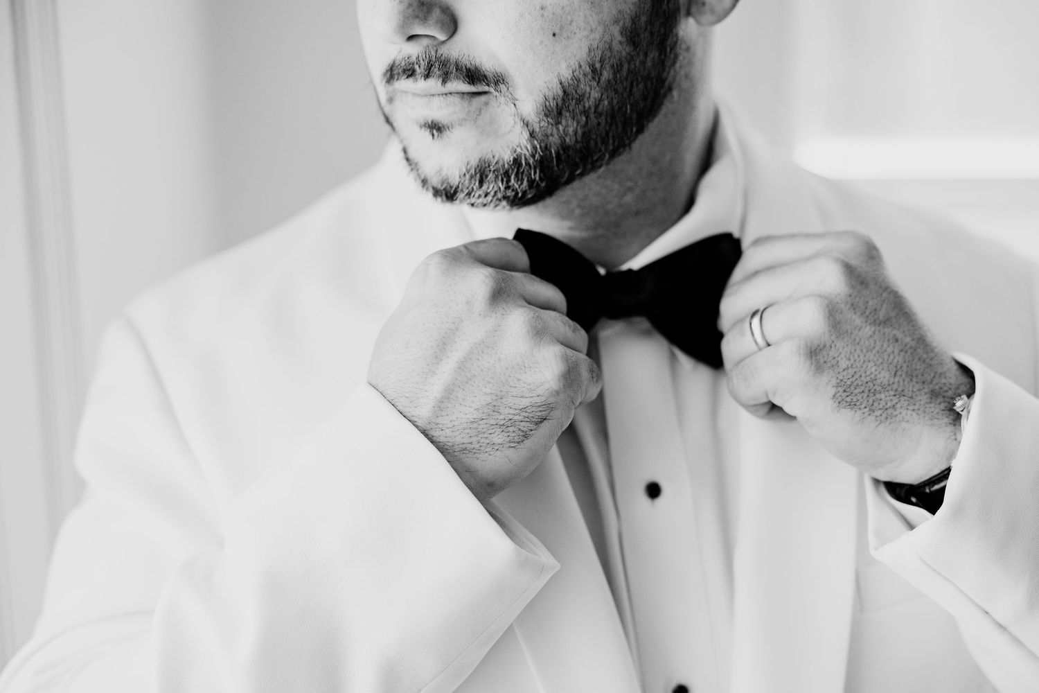 Close-up black and white photo of groom adjusting black bow tie on white tuxedo jacket.