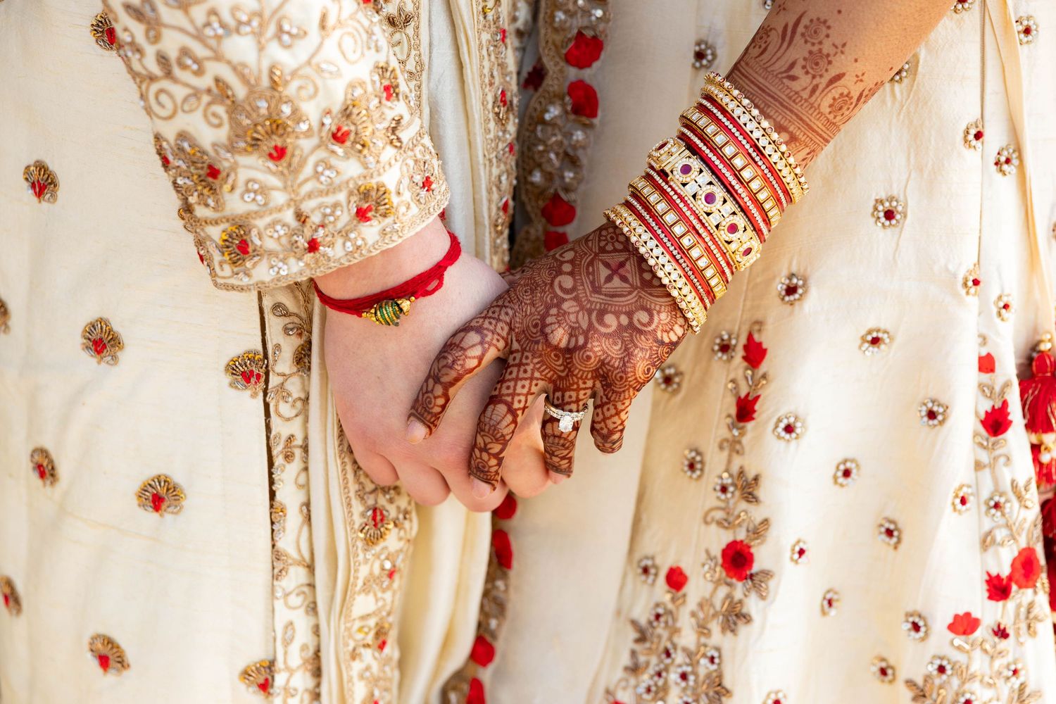 Indian bride and groom holding hands, showcasing mehndi and gold bangles in matching outfits.