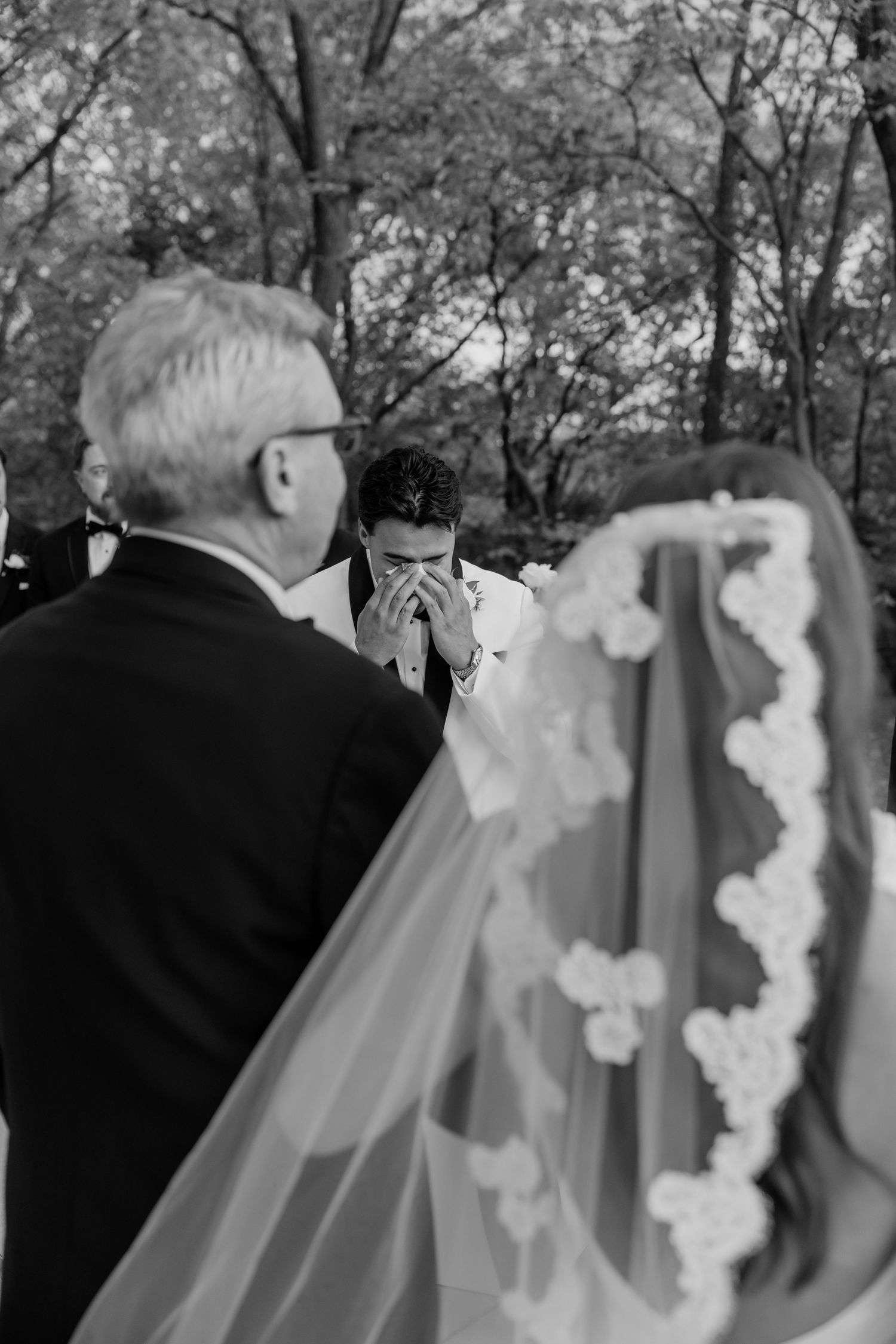 Emotional groom covering face with hands as bride approaches in veil during outdoor wedding ceremony.