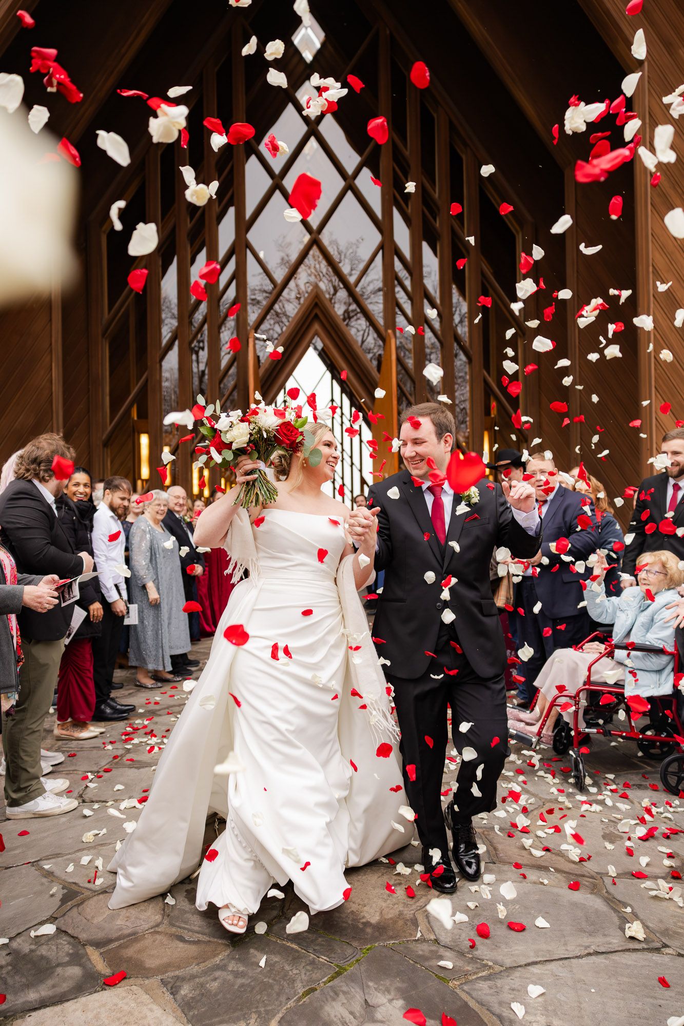 Joyful newlyweds exit church as guests toss red and white rose petals in celebration.
