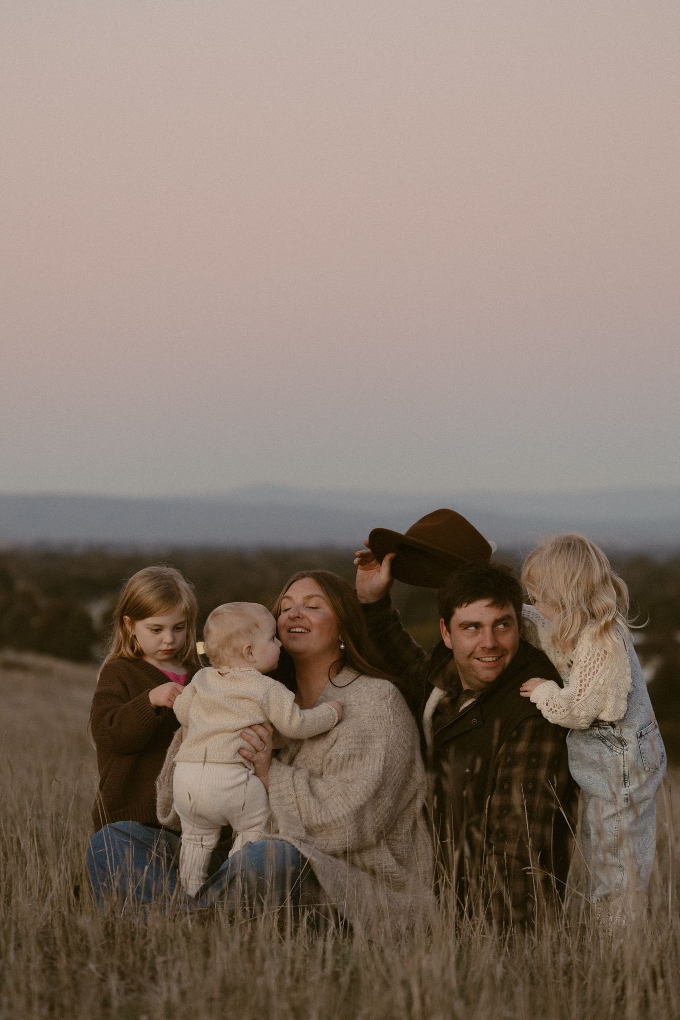 A group of people share laughter and embraces while standing together in a tall grass field at dusk.
