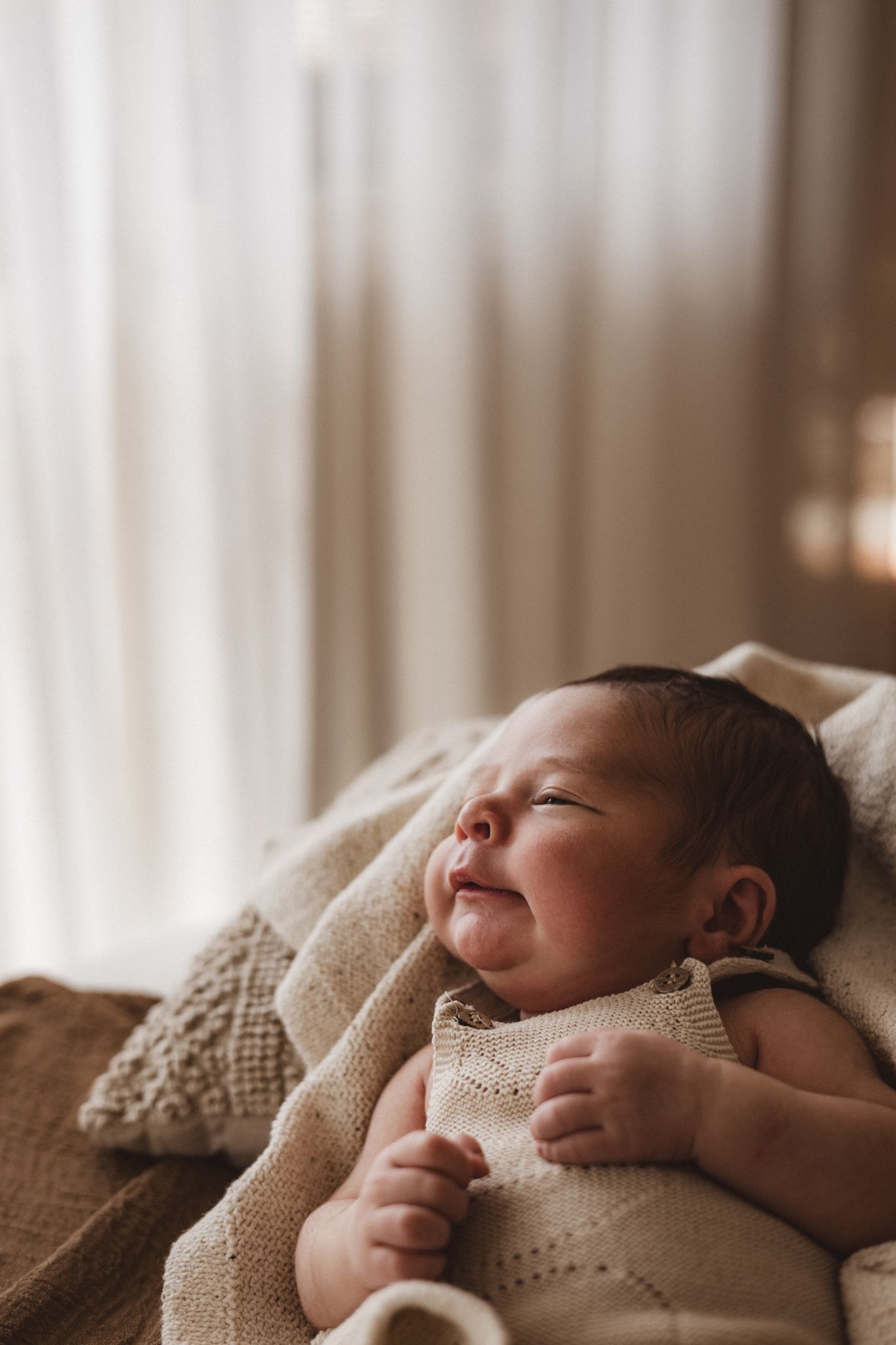 Newborn baby sleeps peacefully under a cozy knit blanket in soft, natural window light.