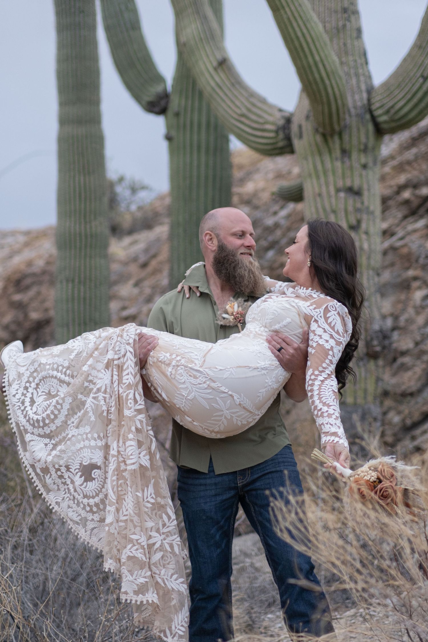 A romantic couple poses in front of tall saguaro cacti in the desert, with the person wearing a flowing white lace dress for an elopement.