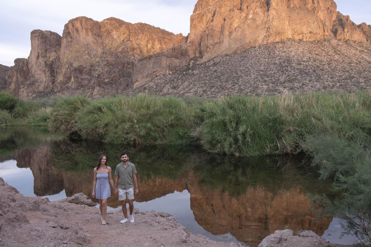 A couple shares an intimate moment near a reflective lake with towering desert cliffs at sunset.