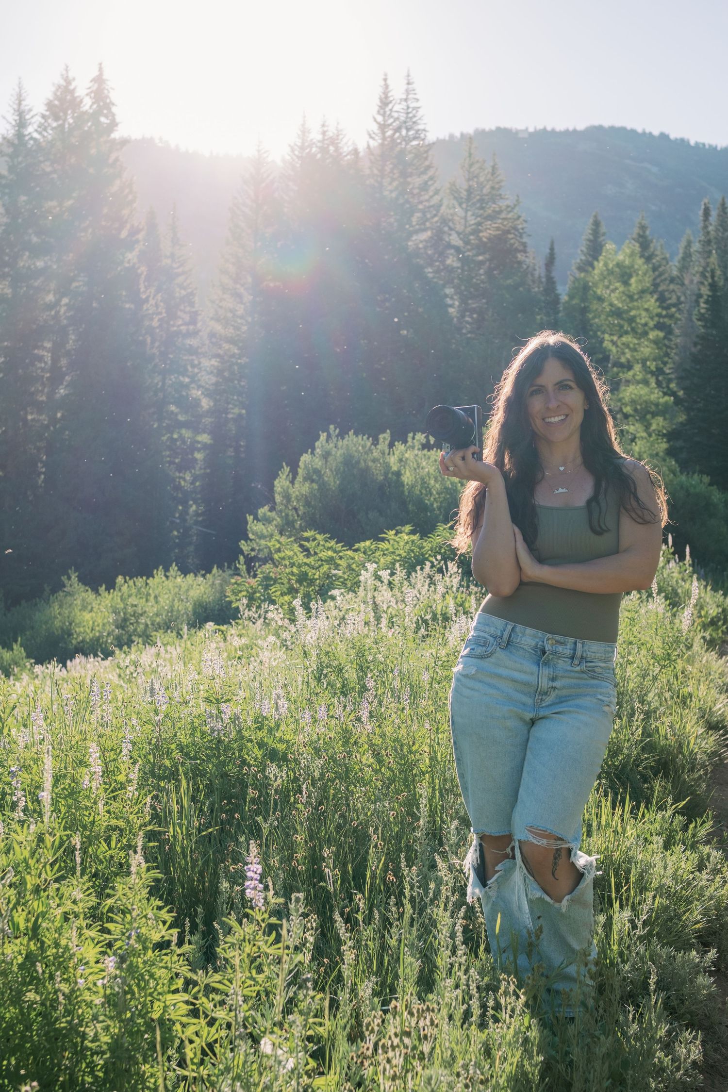 A person enjoying a sunny summer day in a wildflower meadow with evergreen trees and mountains in the backdrop.