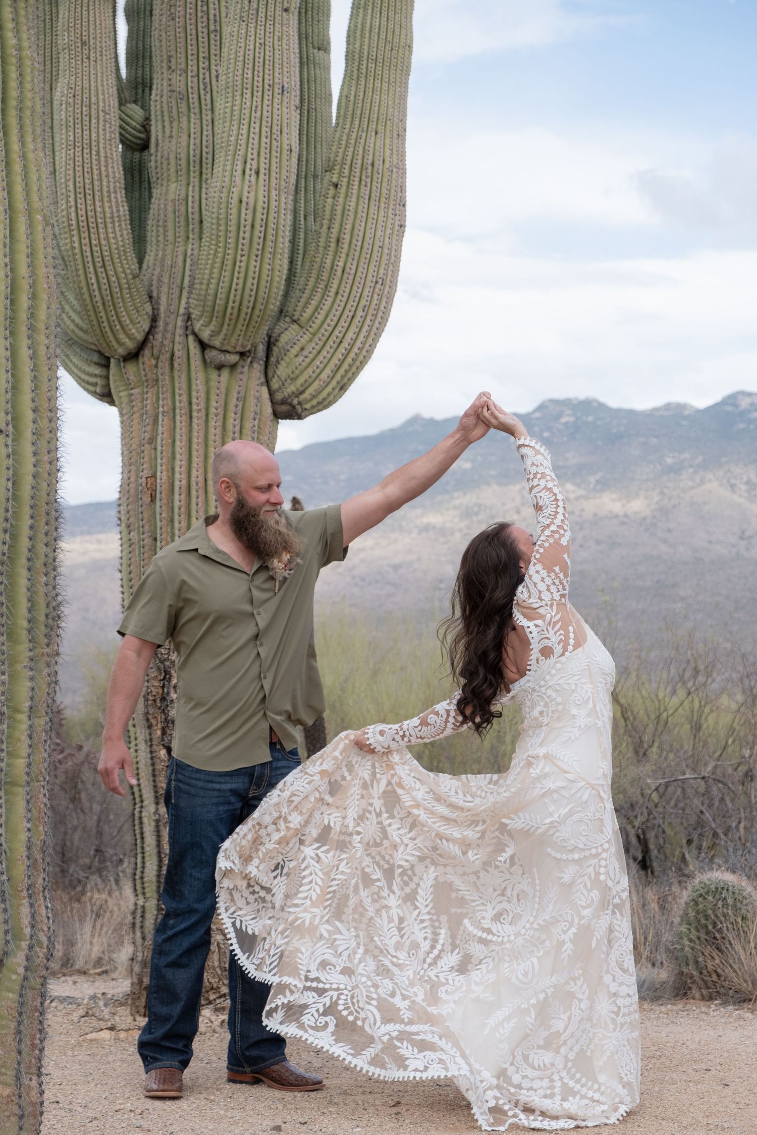 A couple in wedding attire dance together in front of a tall saguaro cactus with desert mountains in the background for their elopement.