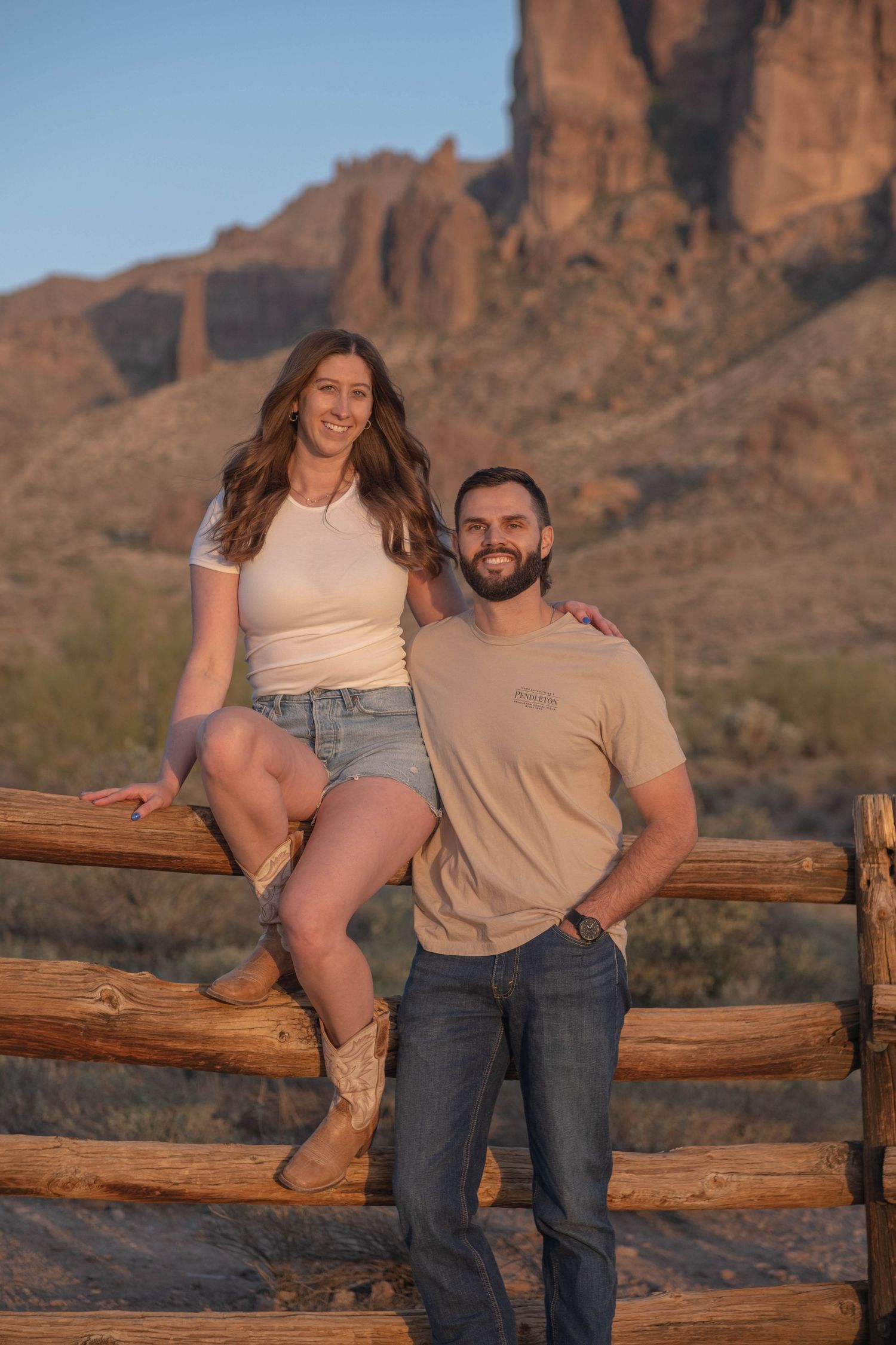 A couple stands by a wooden fence with towering desert cliffs in the background.